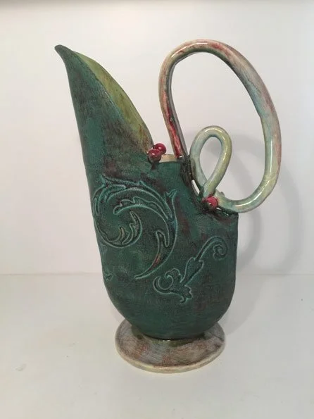 A ceramic pitcher with a curved handle and decorative swirling patterns, featuring a green and brown glaze.