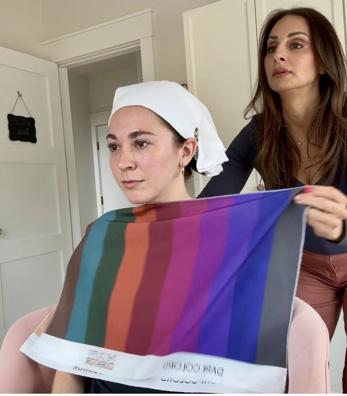 A woman sitting with a colorful test pattern sheet held over her eyes, a woman standing next to her holding the sheet, both in a room with white walls and a door with a WELCOME sign.
