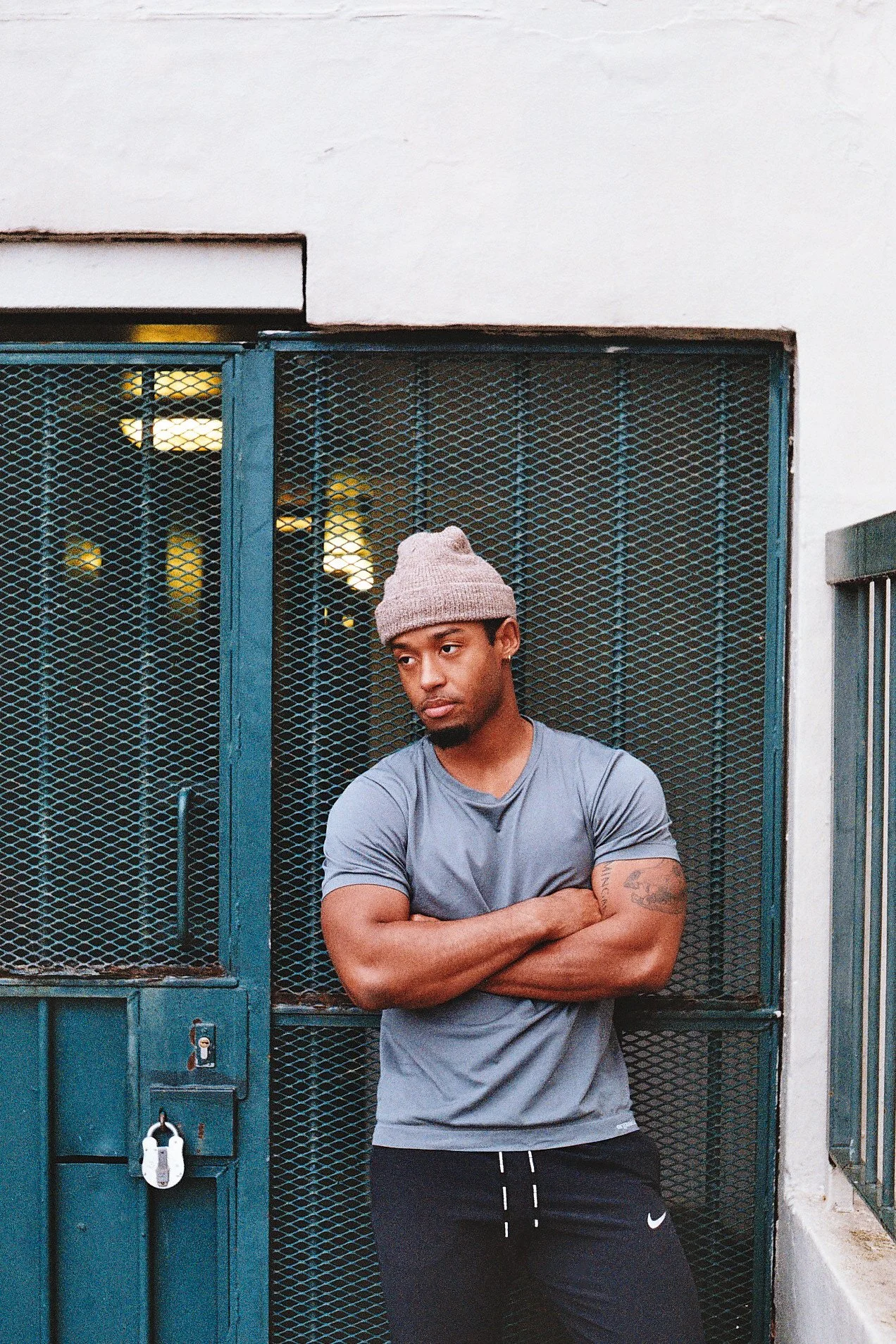 A young man in a gray t-shirt, black shorts, and a beige knit cap, standing with arms crossed in front of a blue metal gate with a lock, in an urban outdoor setting.
