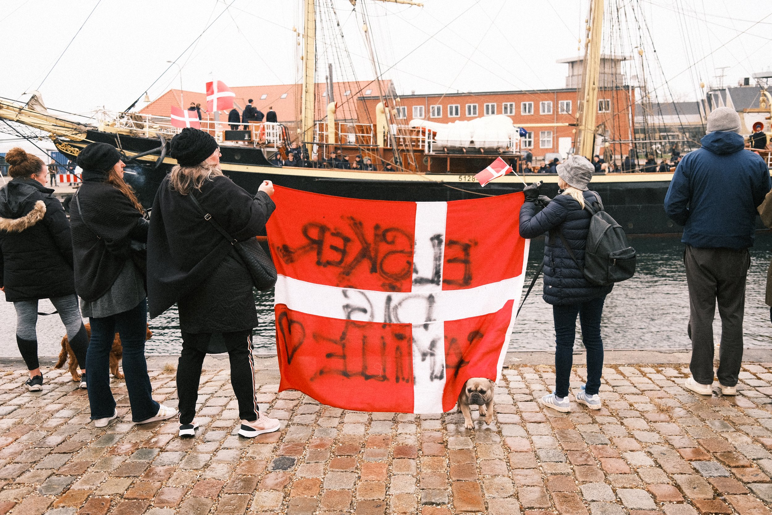 Group of people holding Danish flags gathered near a large ship docked at a harbor with red brick buildings in the background.