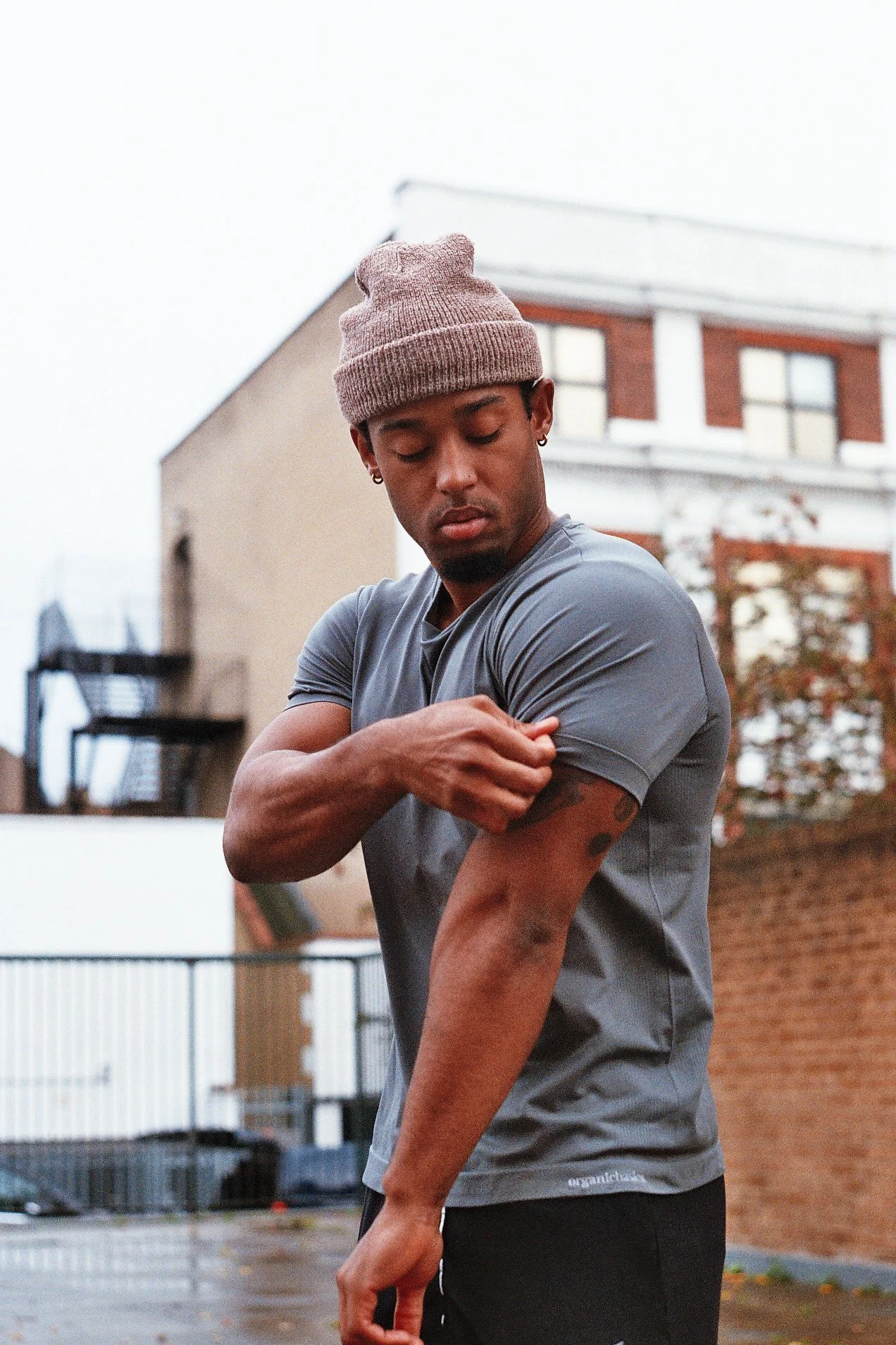 A young man wearing a gray t-shirt and a beige beanie is standing outdoors, rolling up his sleeve to reveal a muscle on his arm. The background shows a brick building with trees and a metal fence.