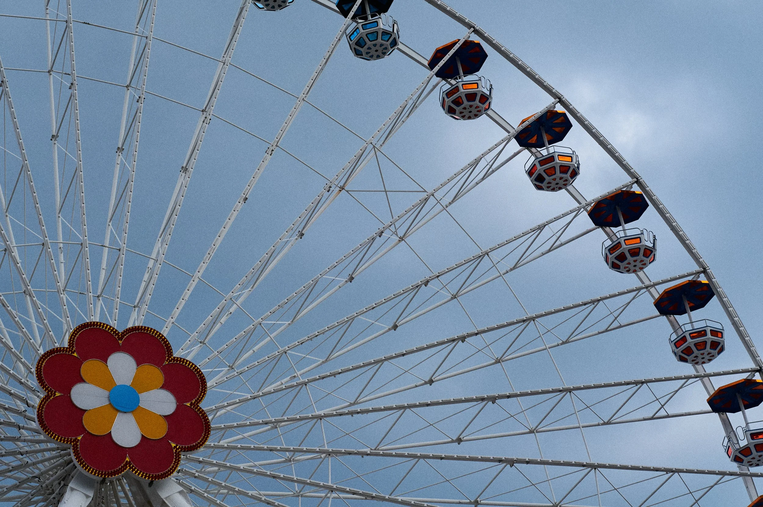 A Ferris wheel with colorful gondolas against a cloudy sky, featuring a large red and yellow flower-shaped decoration at the bottom.