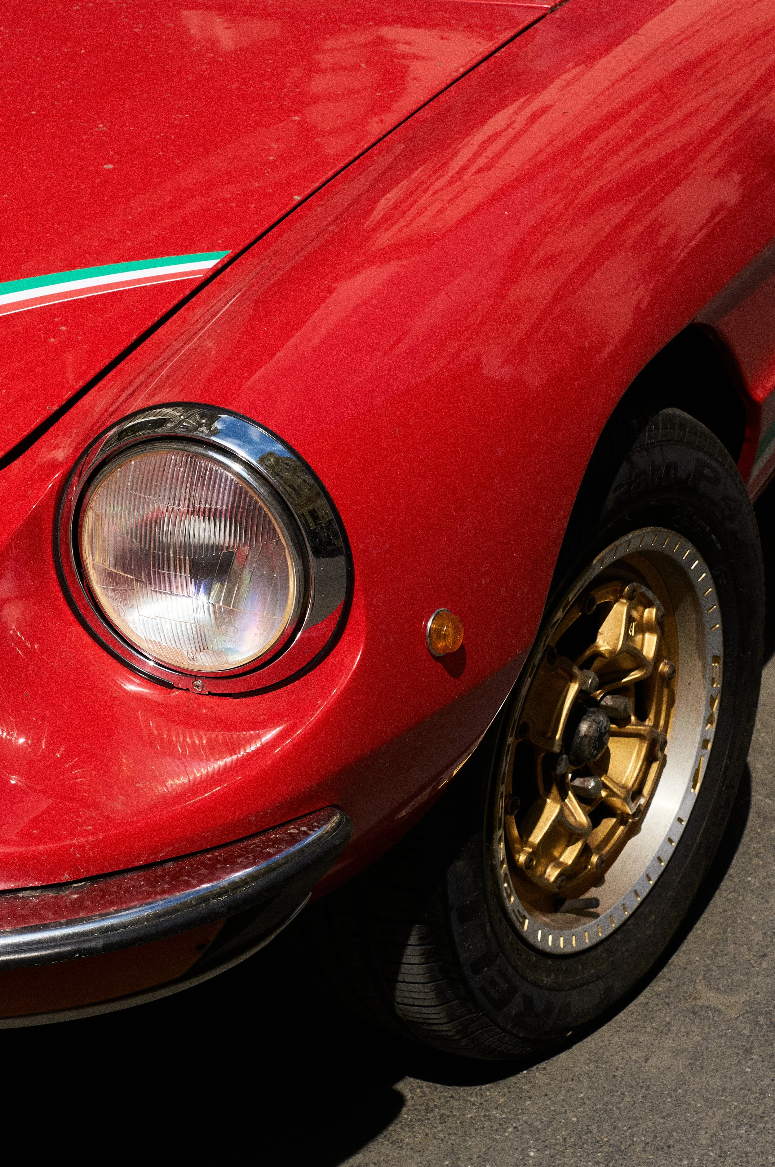 Close-up of the front left side of a red car with a gold wheel and black tire, featuring a headlight, part of the bumper, and a small amber side marker light. The car has a small strip of green, white, and red stripes on the edge of the hood.