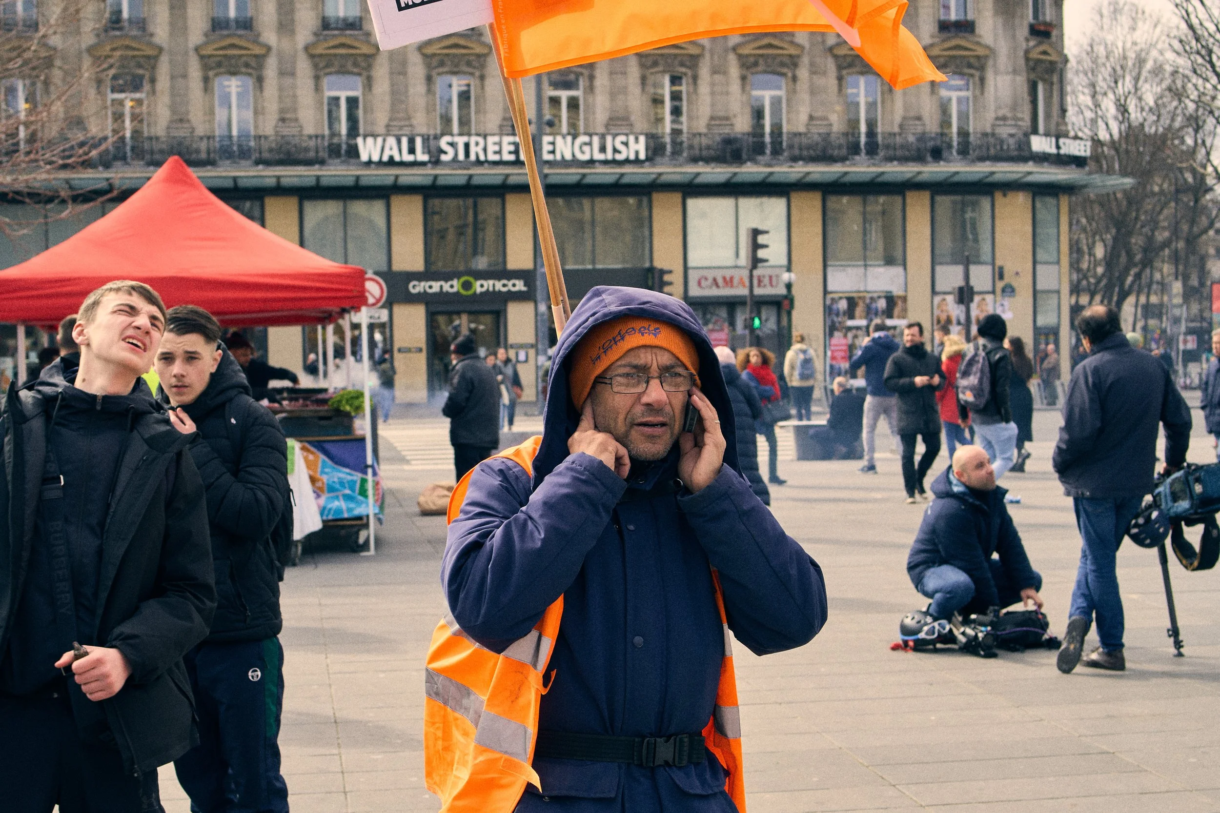 A man wearing glasses, a blue jacket, and an orange safety vest is talking on a cell phone, with a crowd and city buildings in the background during daytime.