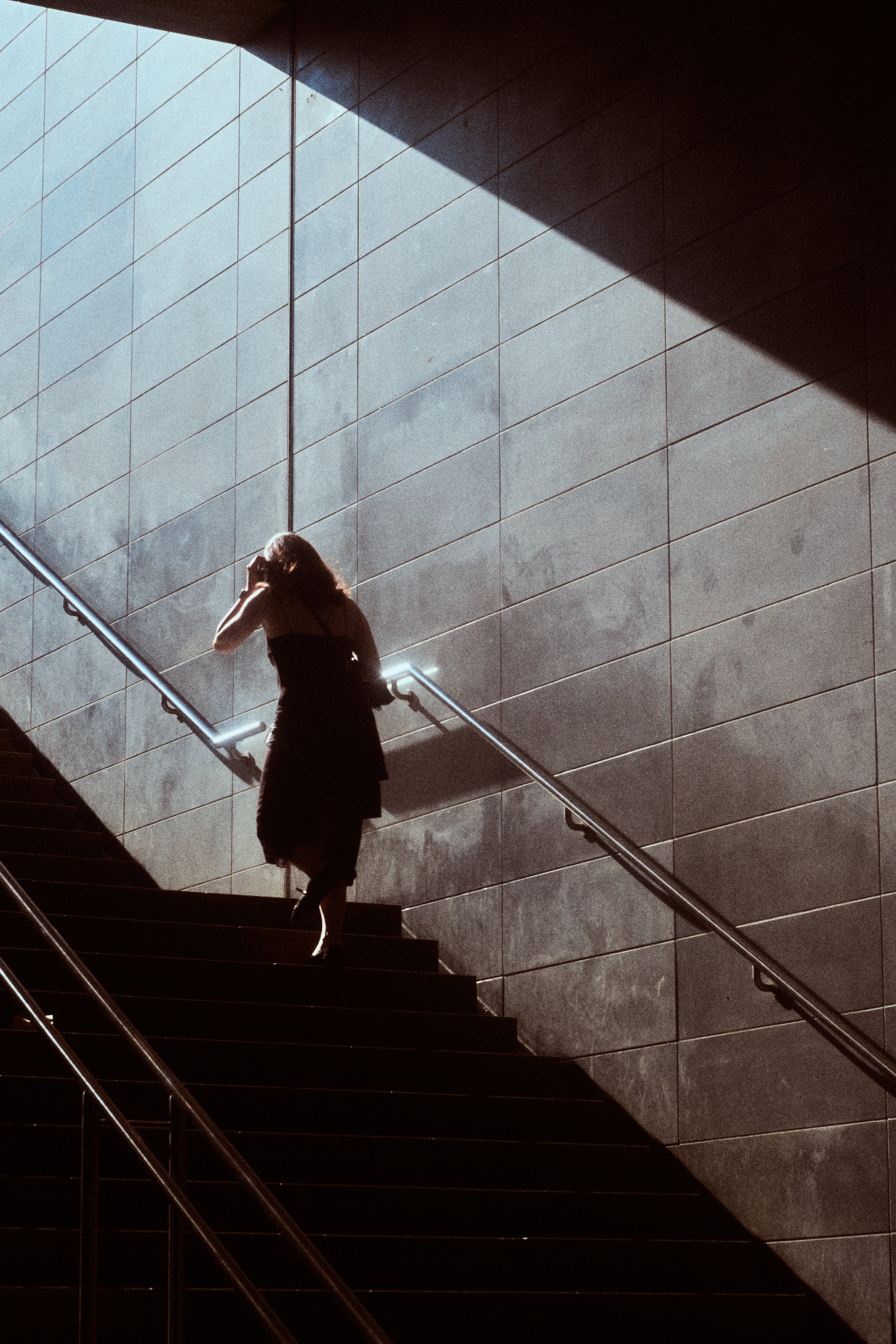A woman with long hair wearing a dress, walking up a staircase with a railing, against a wall made of large rectangular tiles, with sunlight casting a shadow on the wall.