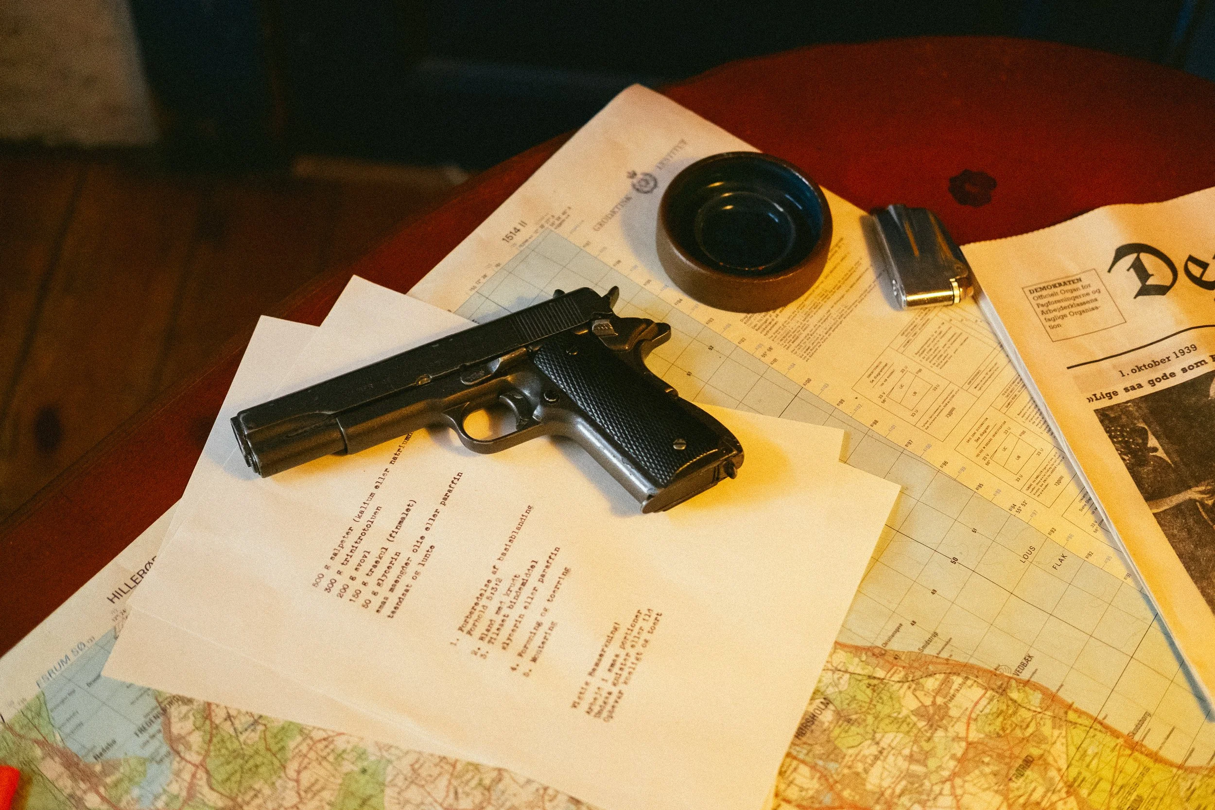 A table with scattered papers, including documents, maps, a black handgun, a roll of black tape, a lighter, and a small file container.