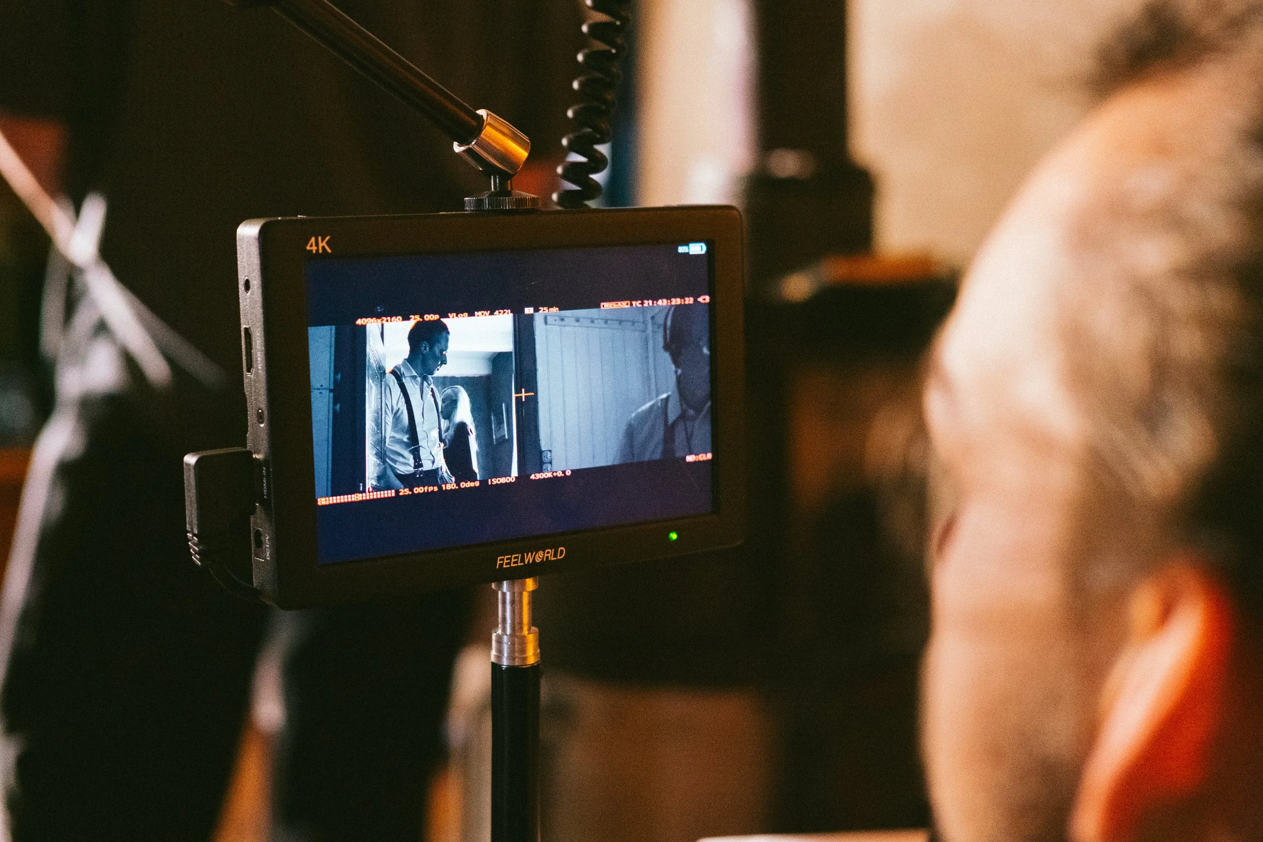 Close-up of a camera monitor on a film set showing a scene with two men, one with a beard and glasses, the other with short hair, engaged in conversation.