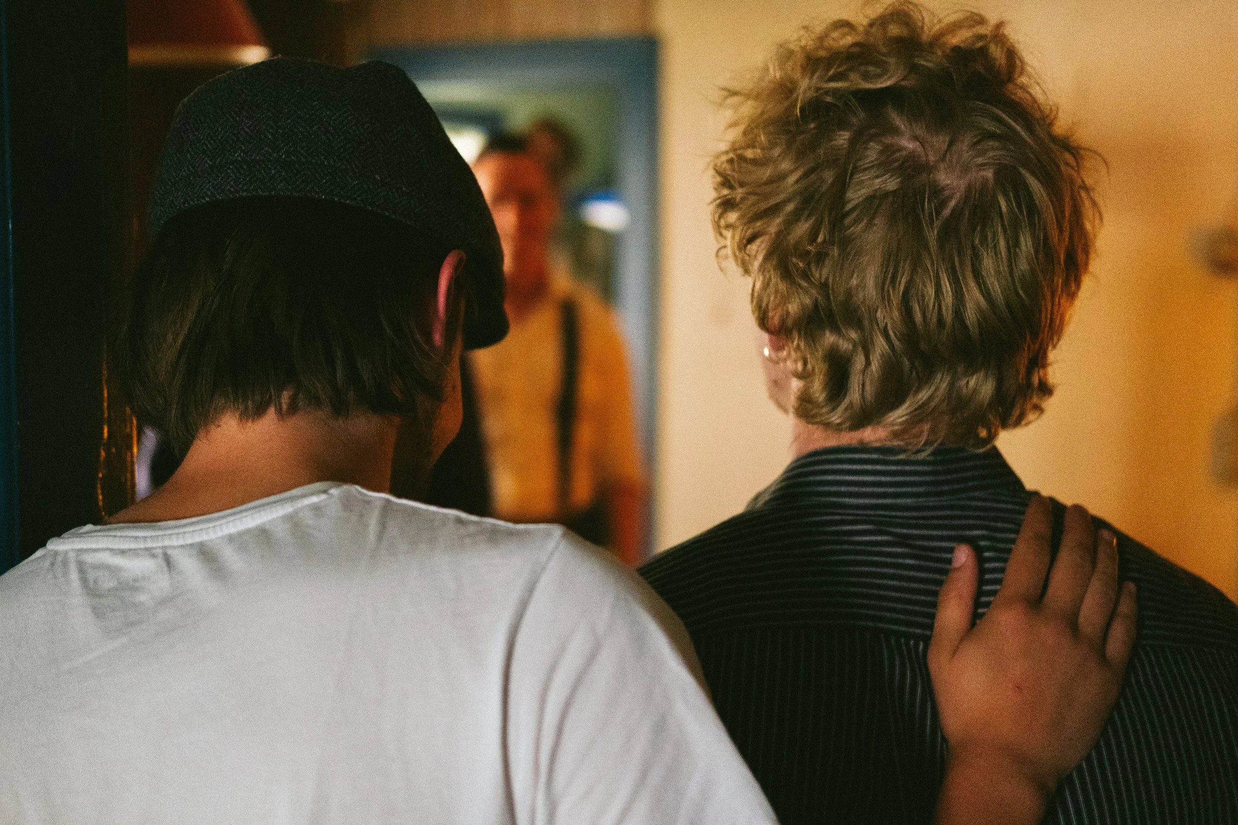Two people with their backs facing the camera; one with short, curly hair in a black and white striped outfit and the other wearing a white top and a dark hat, embracing each other indoors.