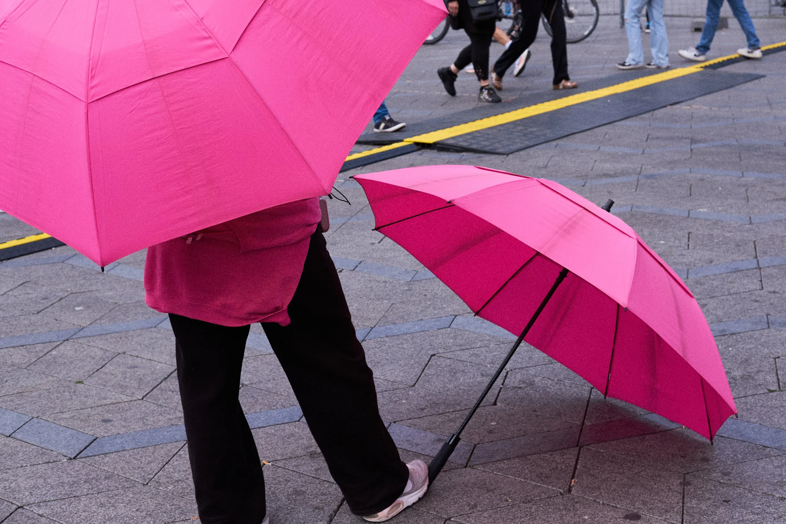 People walking with pink umbrellas on a gray paved street, some people riding bicycles in the background.