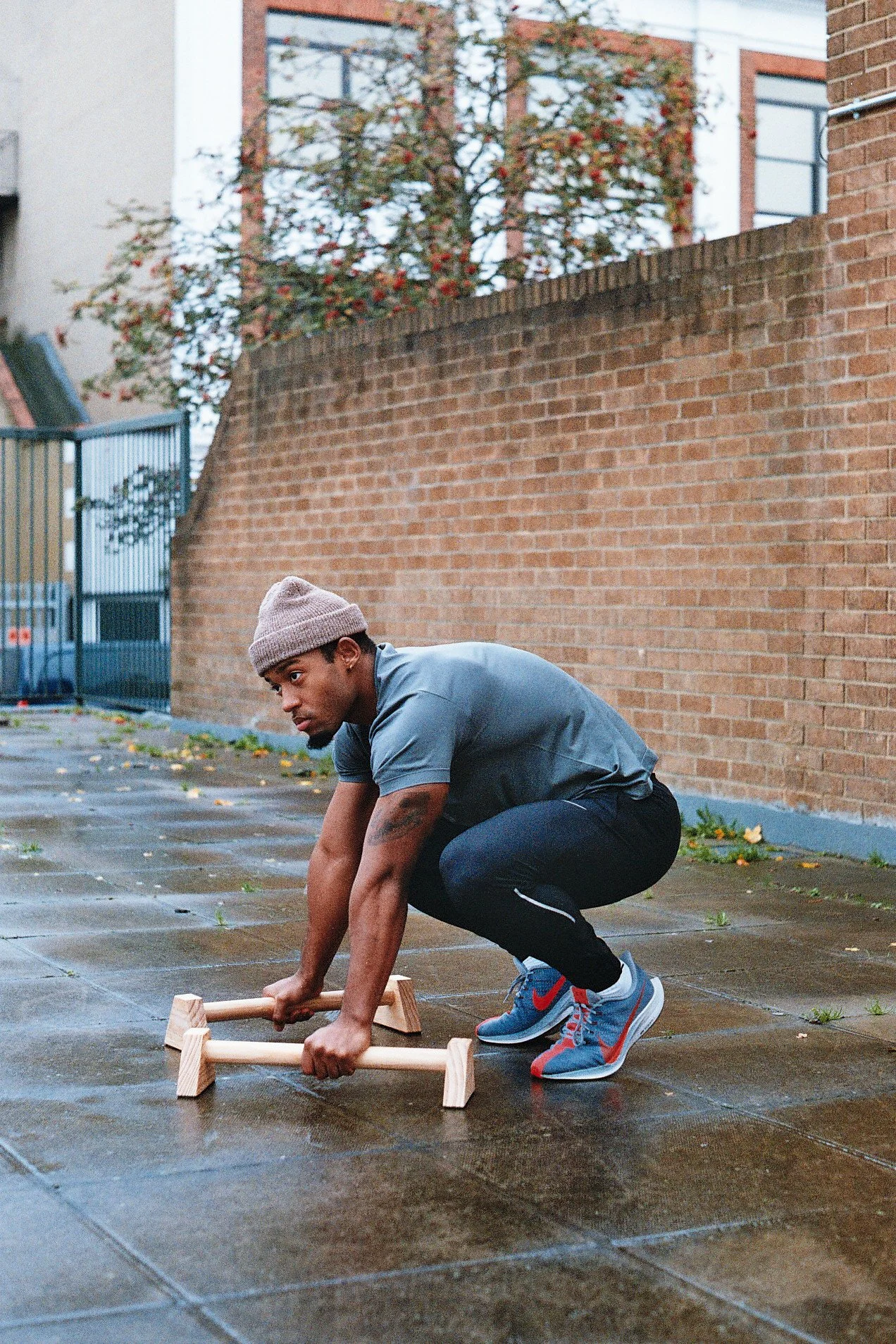 A man wearing a gray beanie, gray t-shirt, black athletic pants, and gray and red sneakers squats on a wet outdoor pavement, holding a small wooden roller with short handles, next to brick walls and a metal fence, with an overcast sky.