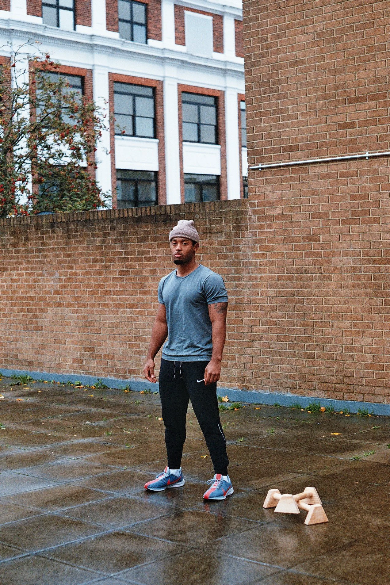 A young man with dark skin, wearing a gray beanie, blue T-shirt, black joggers, and colorful running shoes, standing on a wet outdoor tiled surface next to a brick wall with a pair of wooden workout aids on the ground.