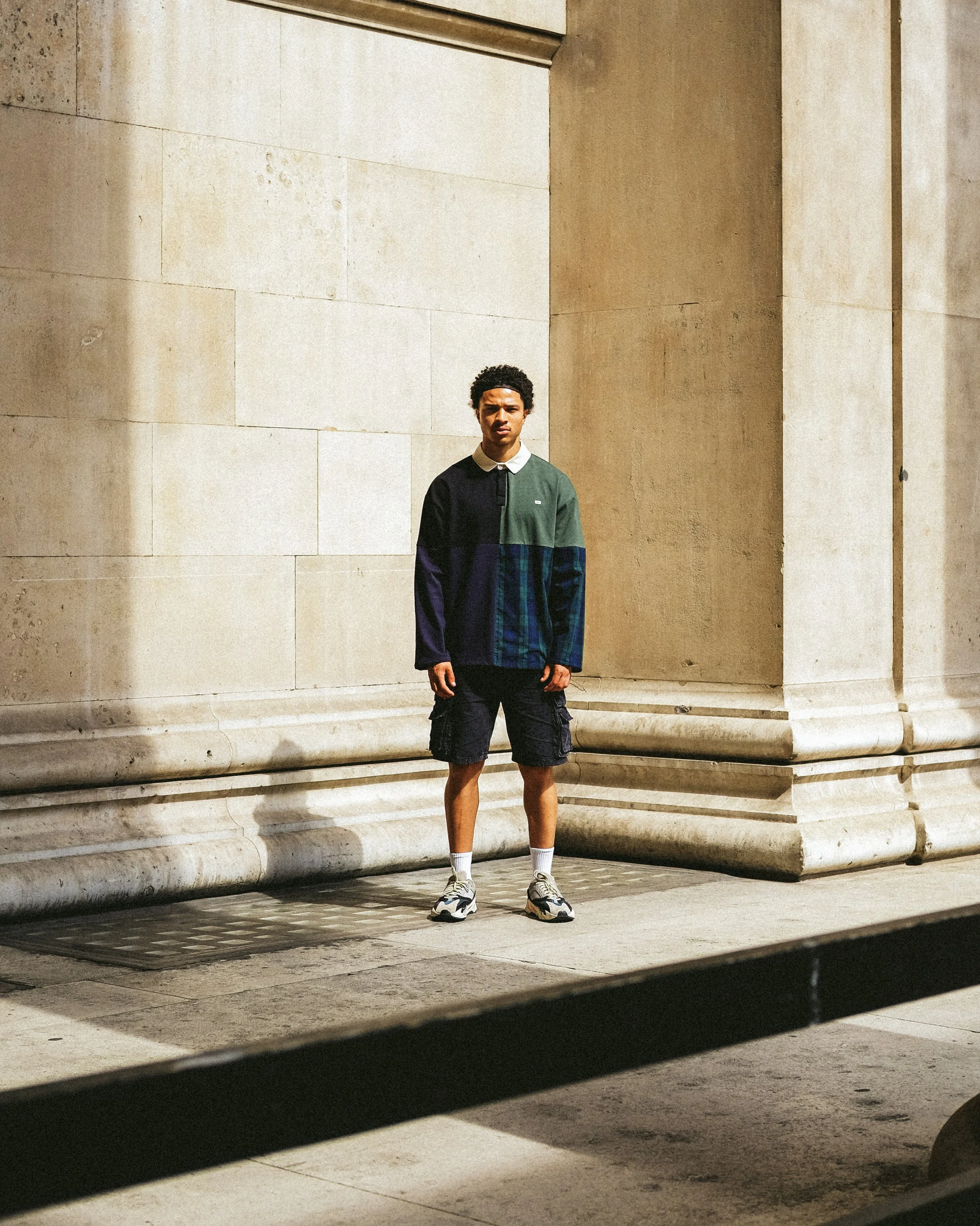 A young man with curly hair, wearing a color-blocked long-sleeve shirt, black shorts, white socks, and sneakers standing against a beige stone wall at night.
