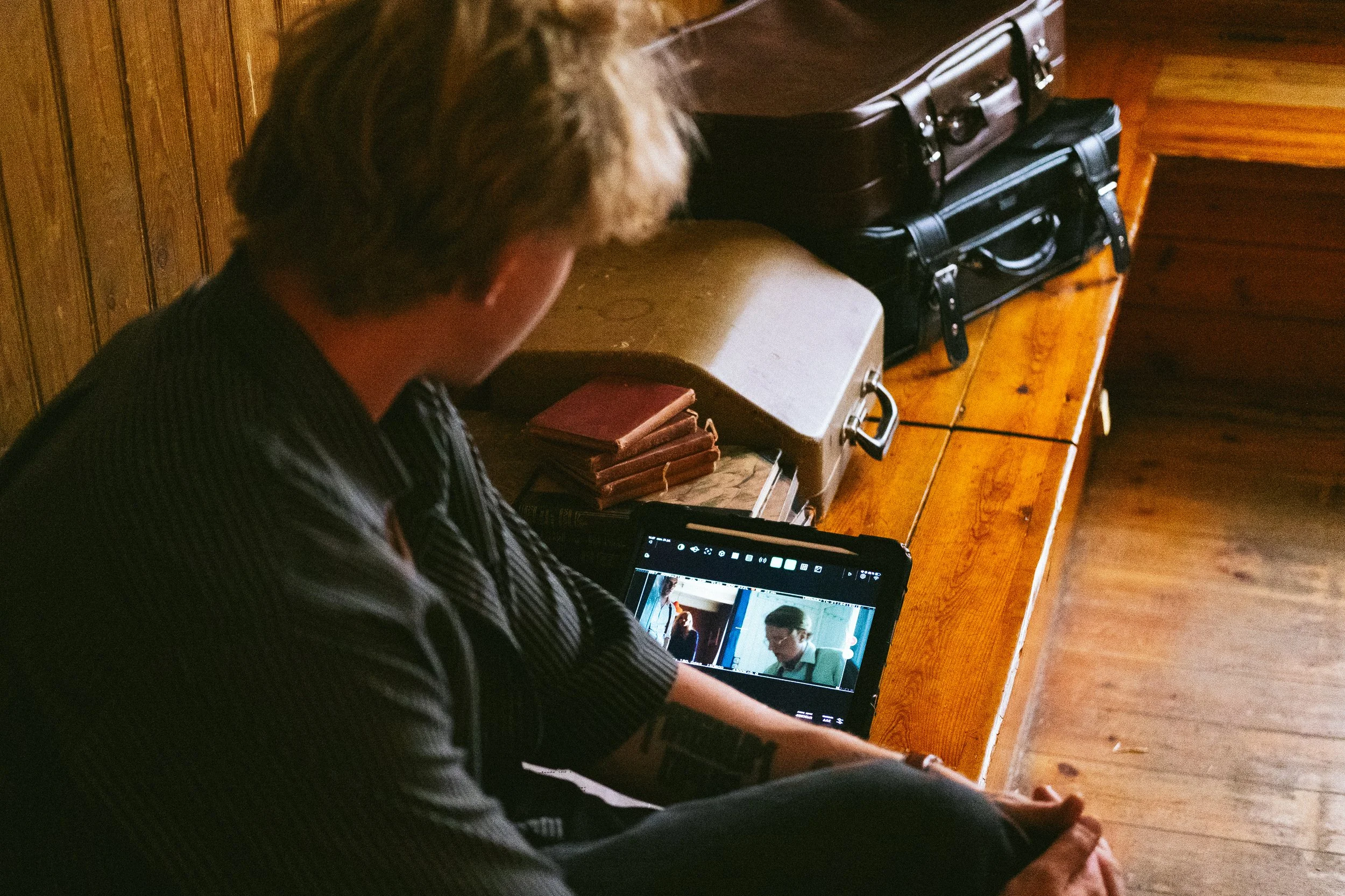 A person with short hair and a striped shirt sitting at a wooden table, looking at a tablet displaying an image of two women having a conversation.