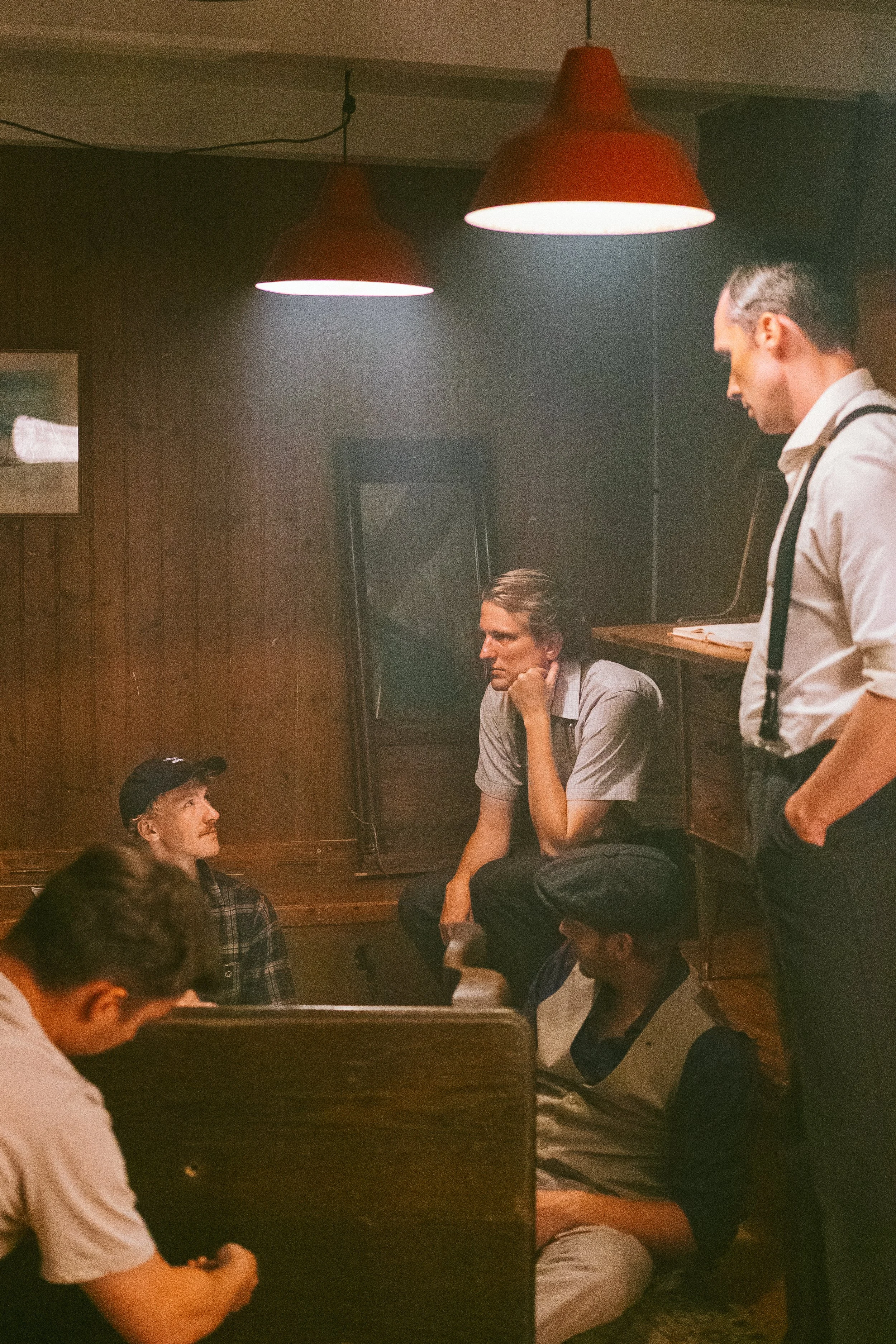 Group of young men in a dimly lit room, some sitting and some standing, engaged in conversation, with warm lighting from overhead lamps.