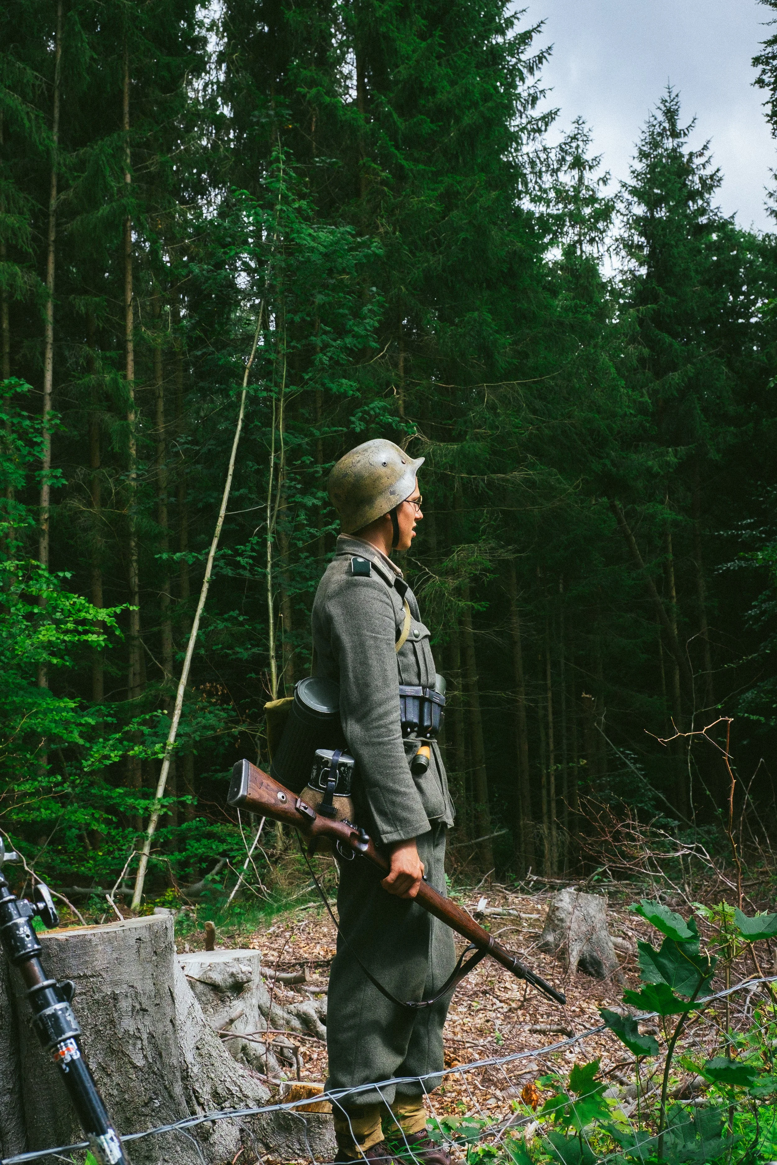 A person dressed in military uniform with a helmet, holding a rifle, standing in a forested area with trees, logs, and a wire fence.