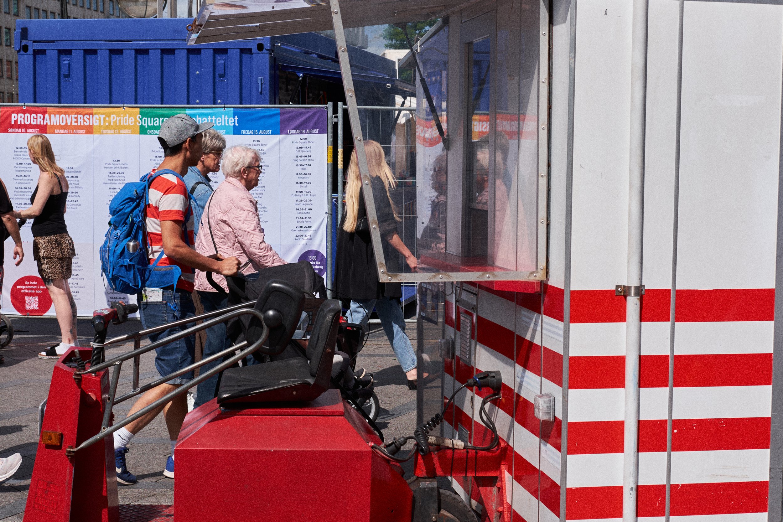 People standing in line at a red and white striped mobile ticket booth in a city square, with a colorful program schedule on a board behind them.