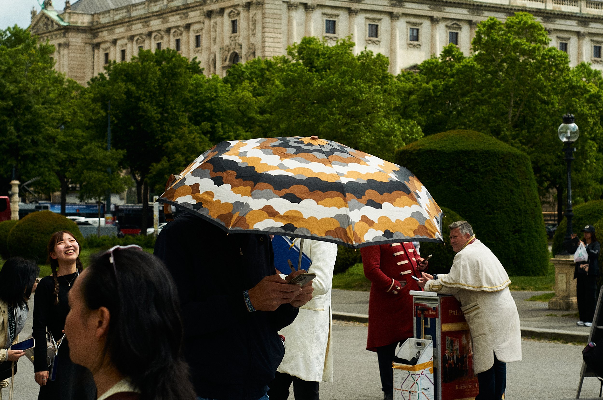 People gathered outdoors near a historic building with lush green trees; one person holds a colorful camouflage umbrella.