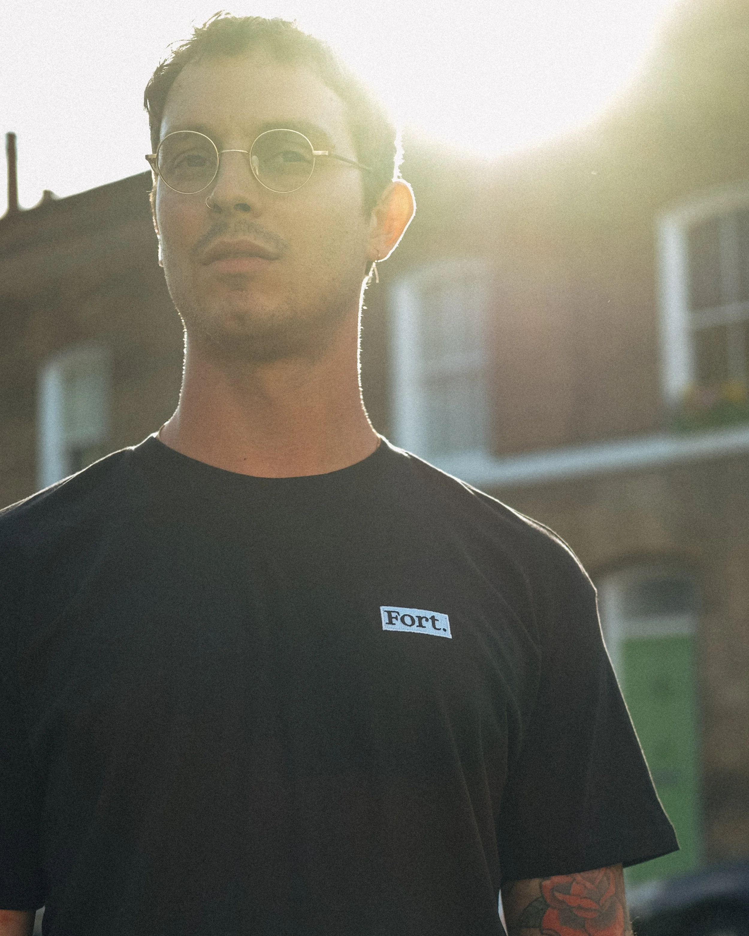 A young man with short dark hair and round sunglasses stands outside in front of a building, with bright sunlight behind him. He is wearing a black t-shirt with a small label that says 'Fort.' on the chest, and has a colorful tattoo sleeve on his lef