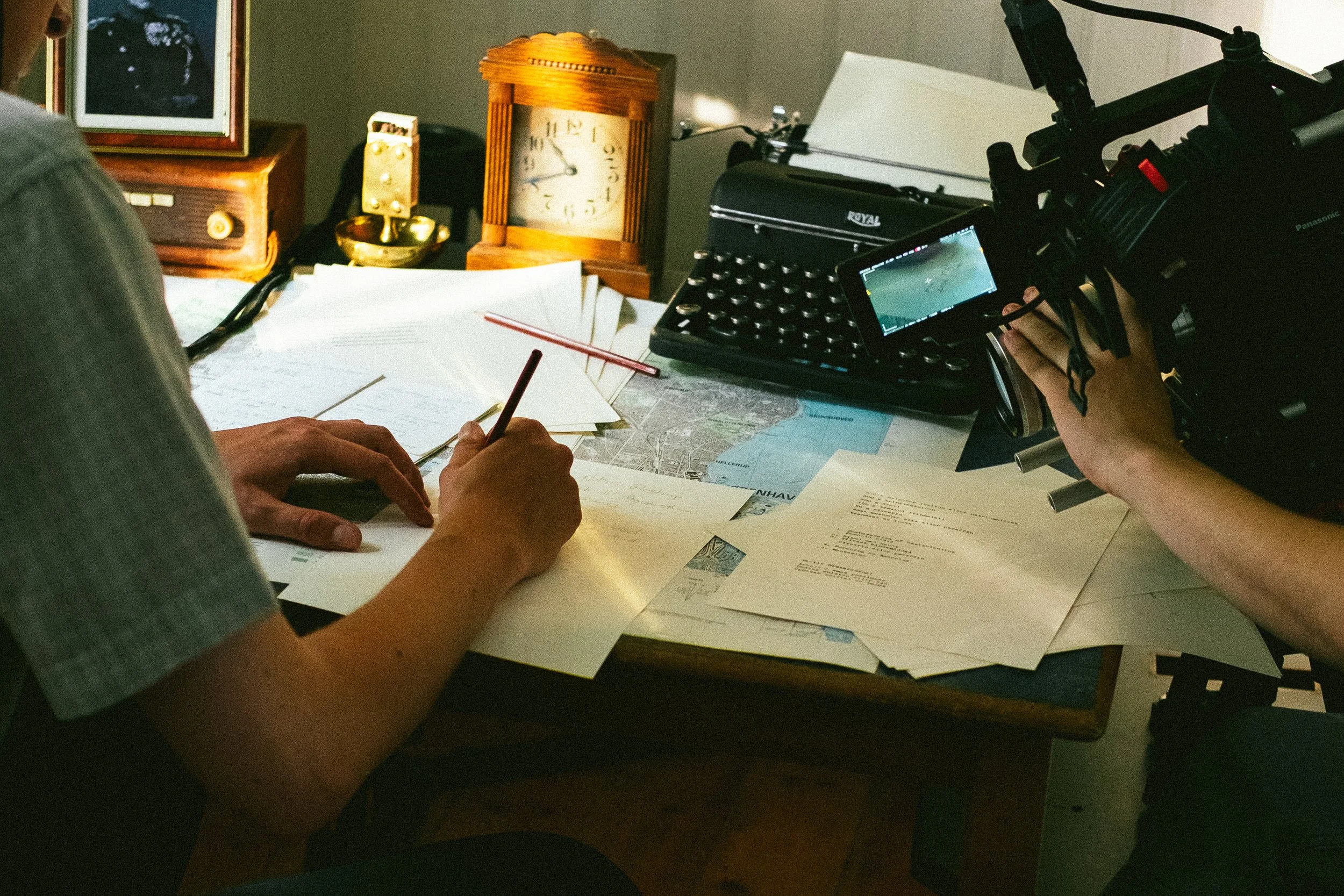 Person writing at a desk with papers, a map, and a pen, with a camera and clocks on the background.