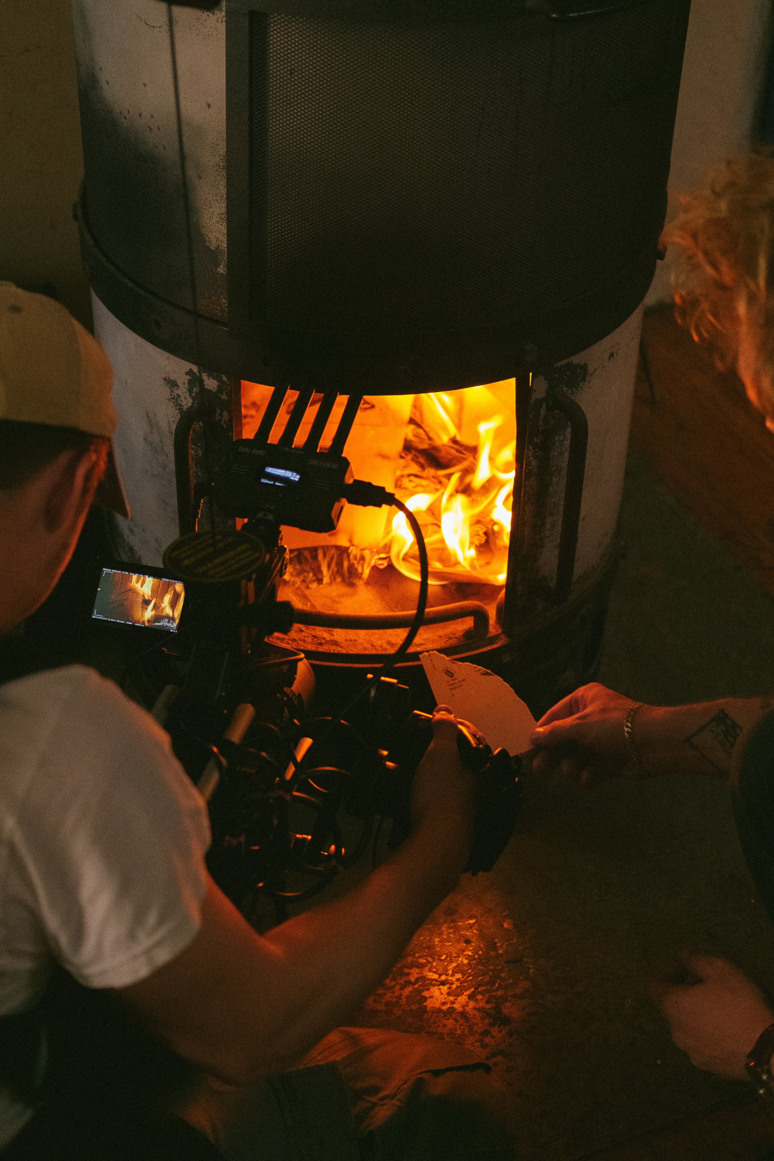 People sitting around a fire pit at night, with some holding papers and equipment, watching the flames.
