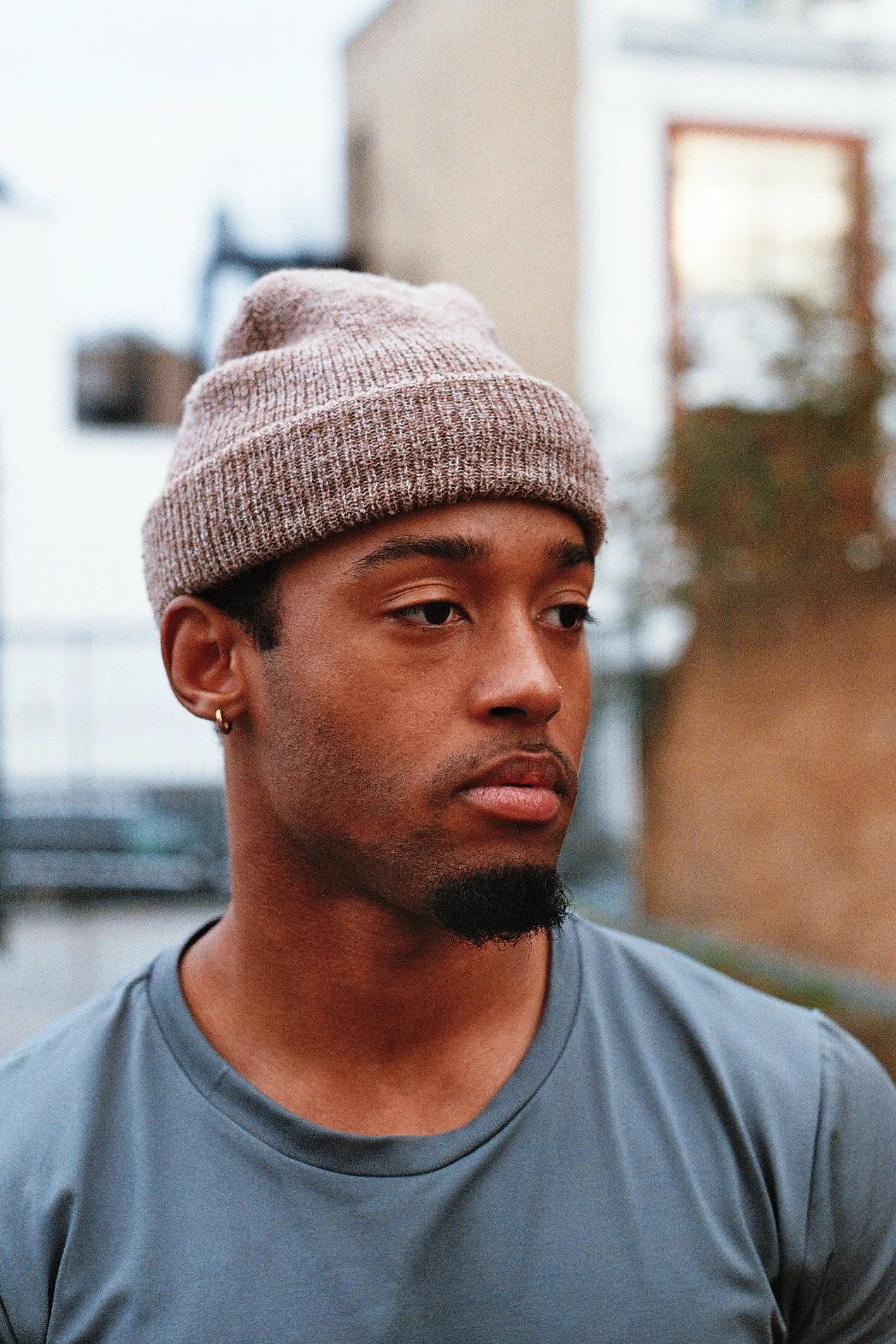 A young man with medium brown skin, a goatee, and mustache, wearing a gray beanie and a gray t-shirt, looking to the side outdoors with blurred background of a house and trees.