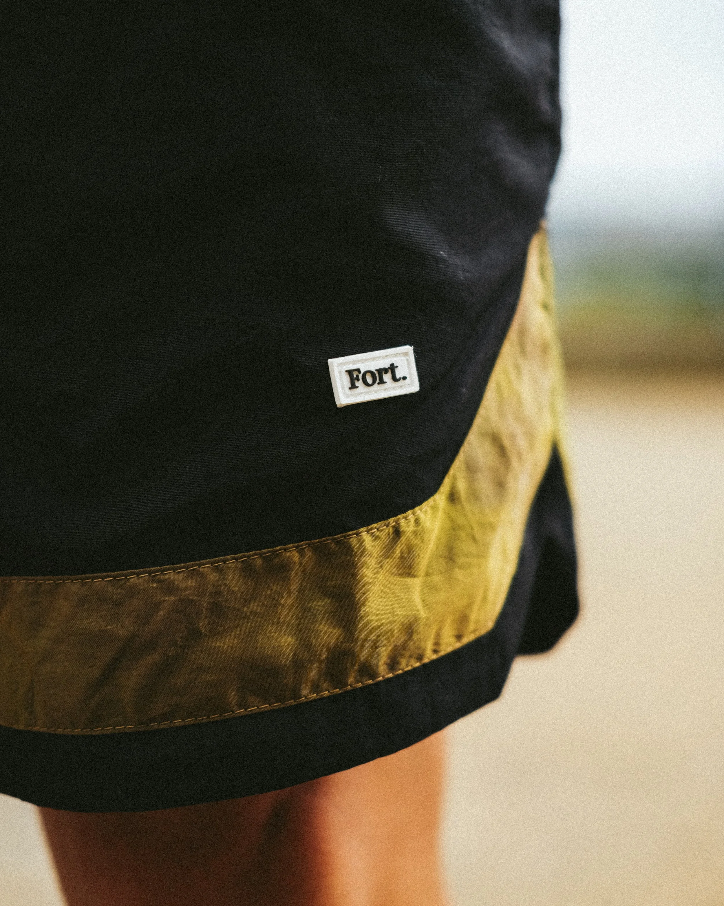 Close-up of a person's leg in shorts, showing a black backpack with a yellow section and a small white label that says 'Fort.'