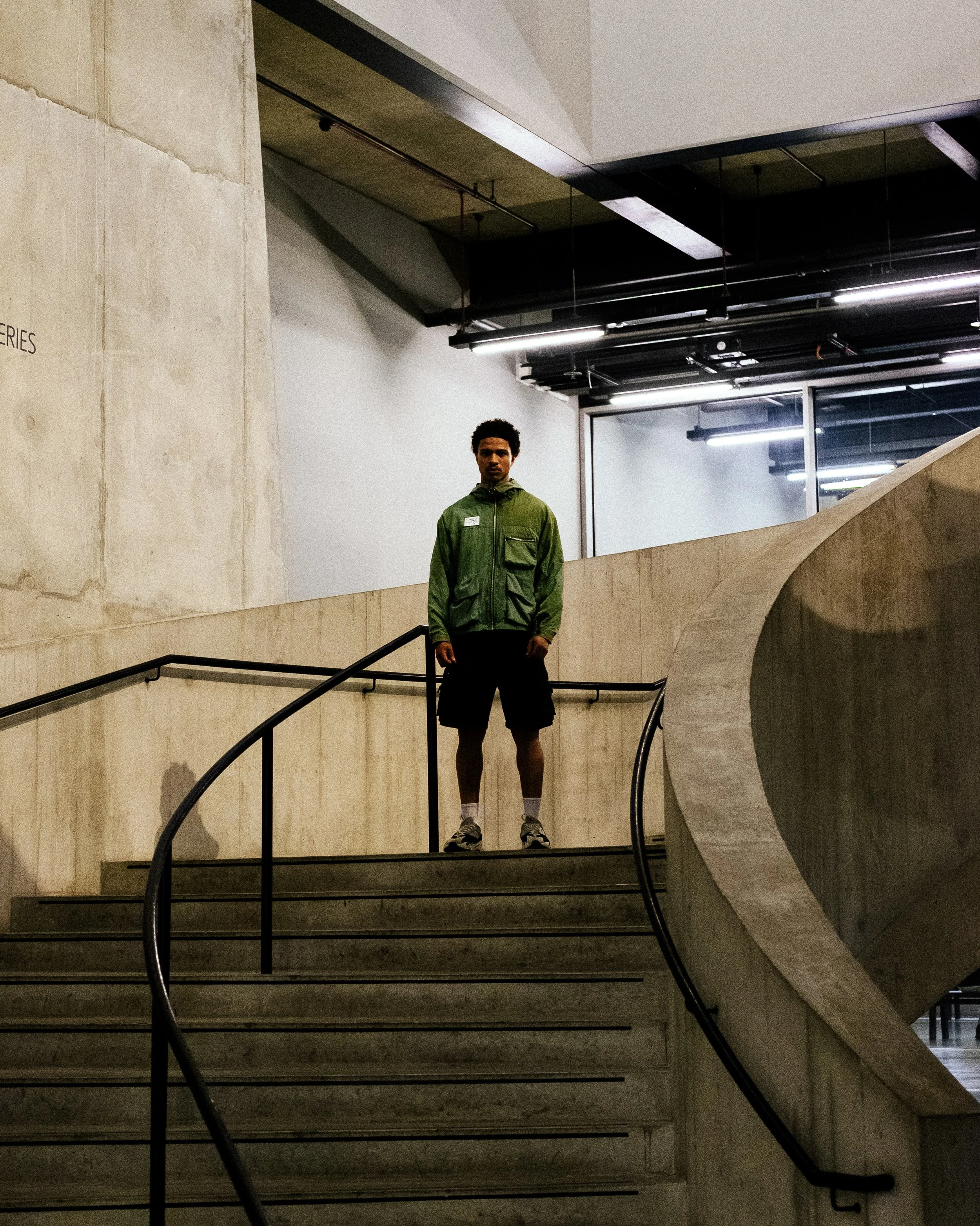A young man standing on a staircase indoors, wearing a green jacket, black shorts, and sneakers, with a serious expression.