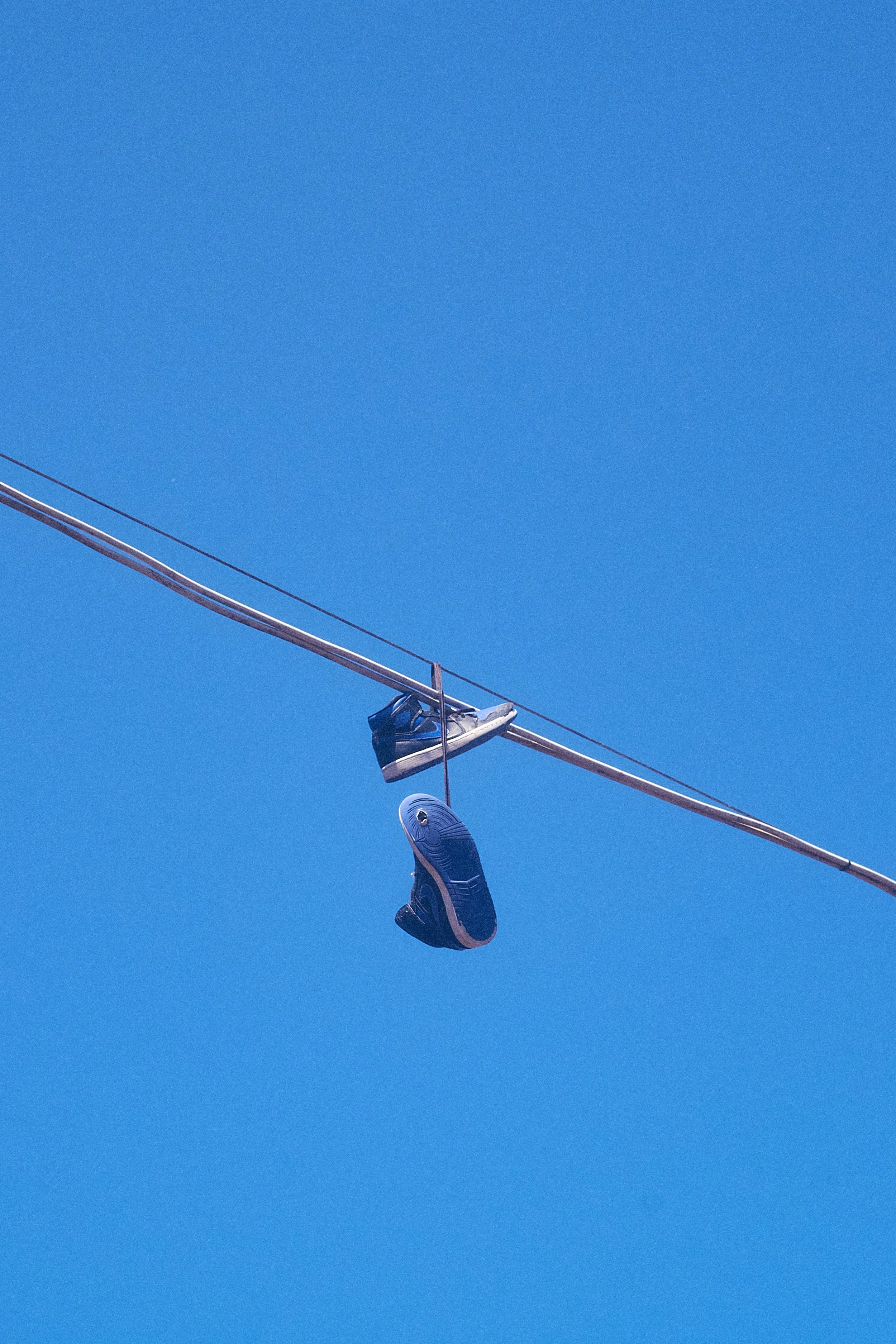 Two athletic shoes hanging from power lines against a clear blue sky.