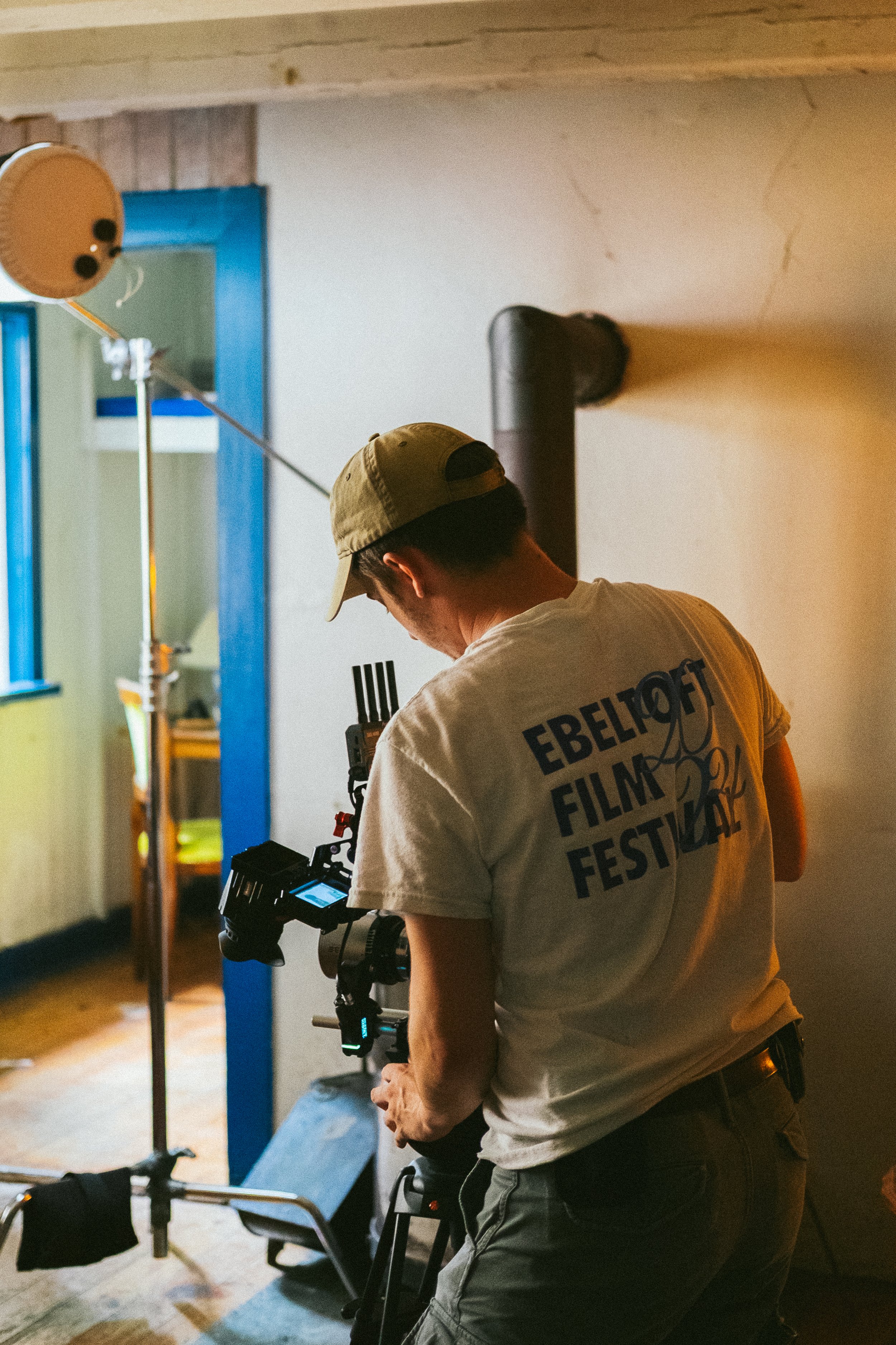 A person wearing a beige cap and a T-shirt with the words 'EBELTOF FILM FESTIVAL' printed on the back, operating a professional camera on a sturdy tripod, with film equipment and light stands set up indoors, possibly on a film set or during a filming