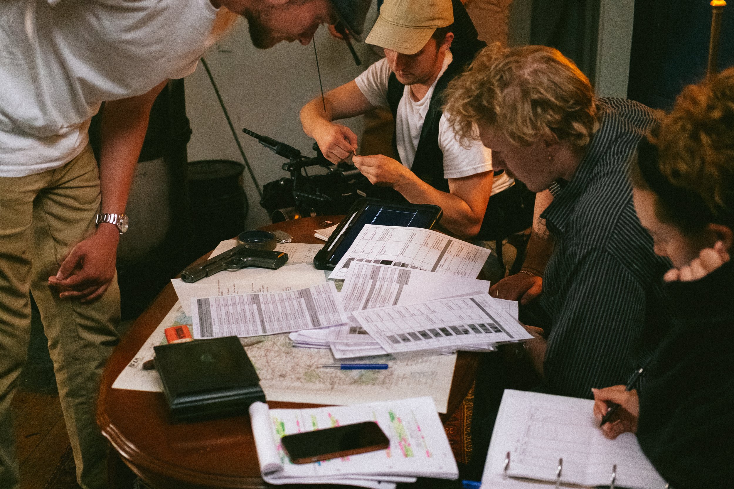 A group of people gathered around a table inspecting documents, with some electronic devices like tablets and smartphones, in an indoor setting.