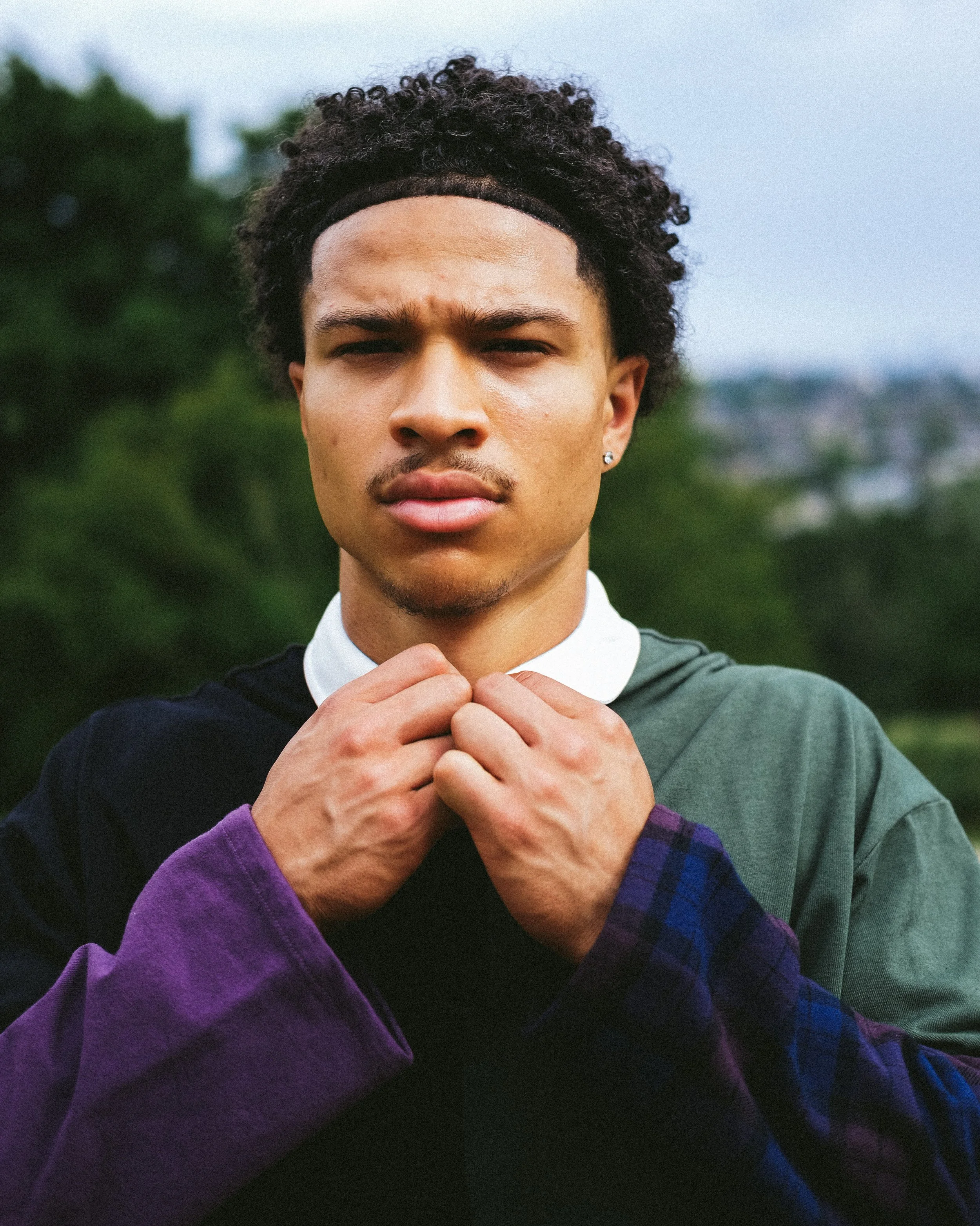 Close-up of a young man with curly hair wearing a dark headband, a green shirt with a white collar, and a purple and plaid sleeve, standing outdoors with a blurred green background.