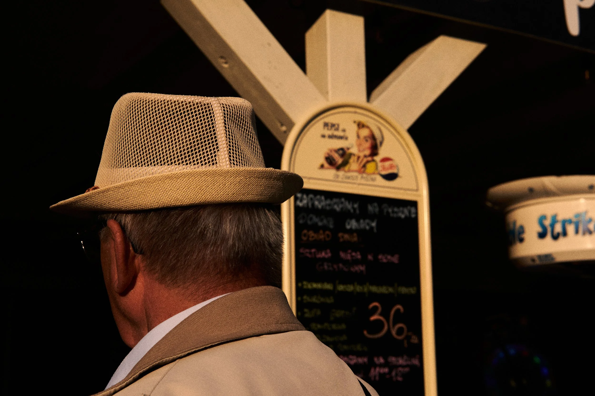 A person wearing a beige fedora hat and a beige suit, seen from behind, standing in front of a menu board at a restaurant or cafe.