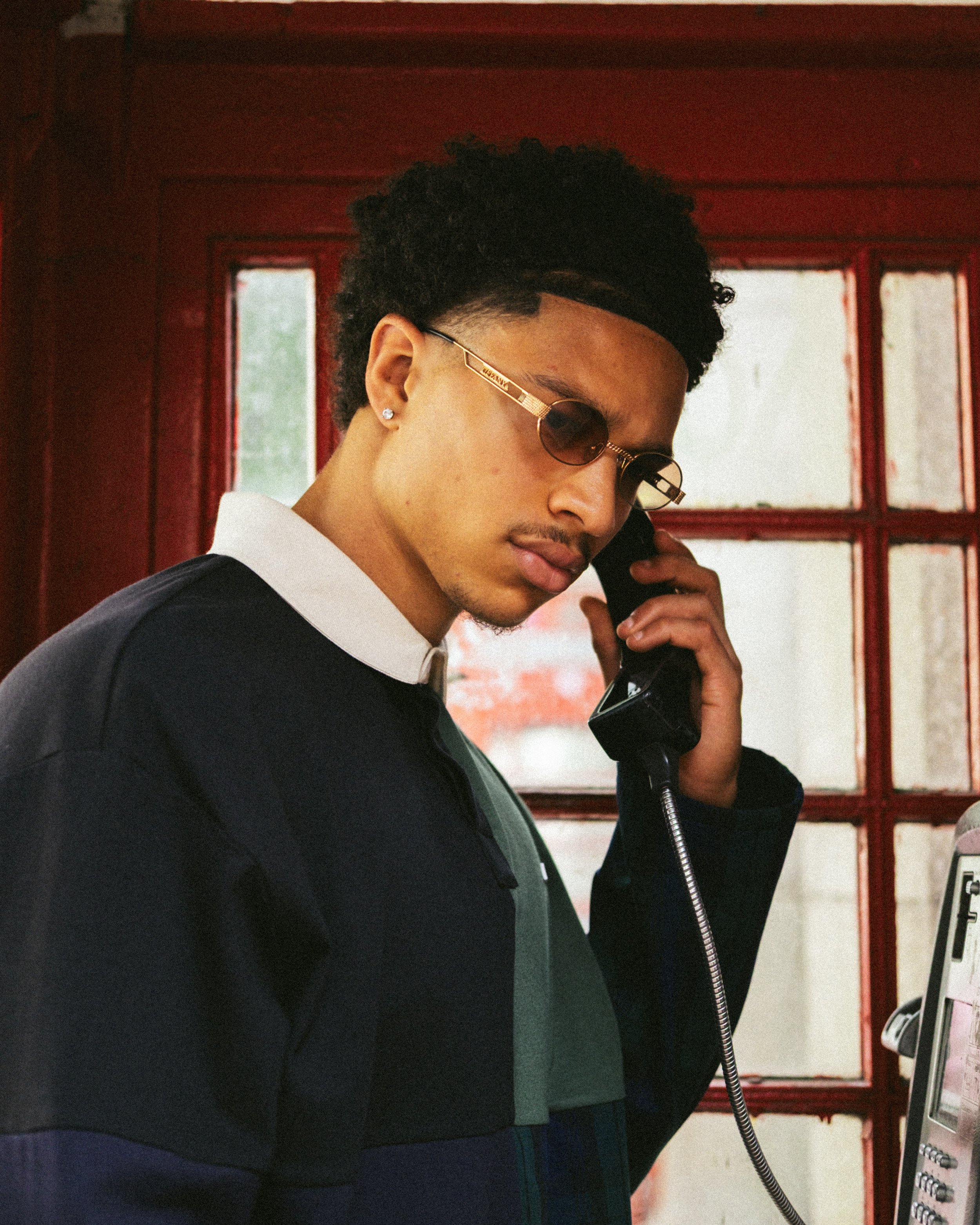 Young man with dark curly hair, wearing sunglasses and a black shirt with a white collar, talking on a payphone inside a red-painted room with windows in the background.