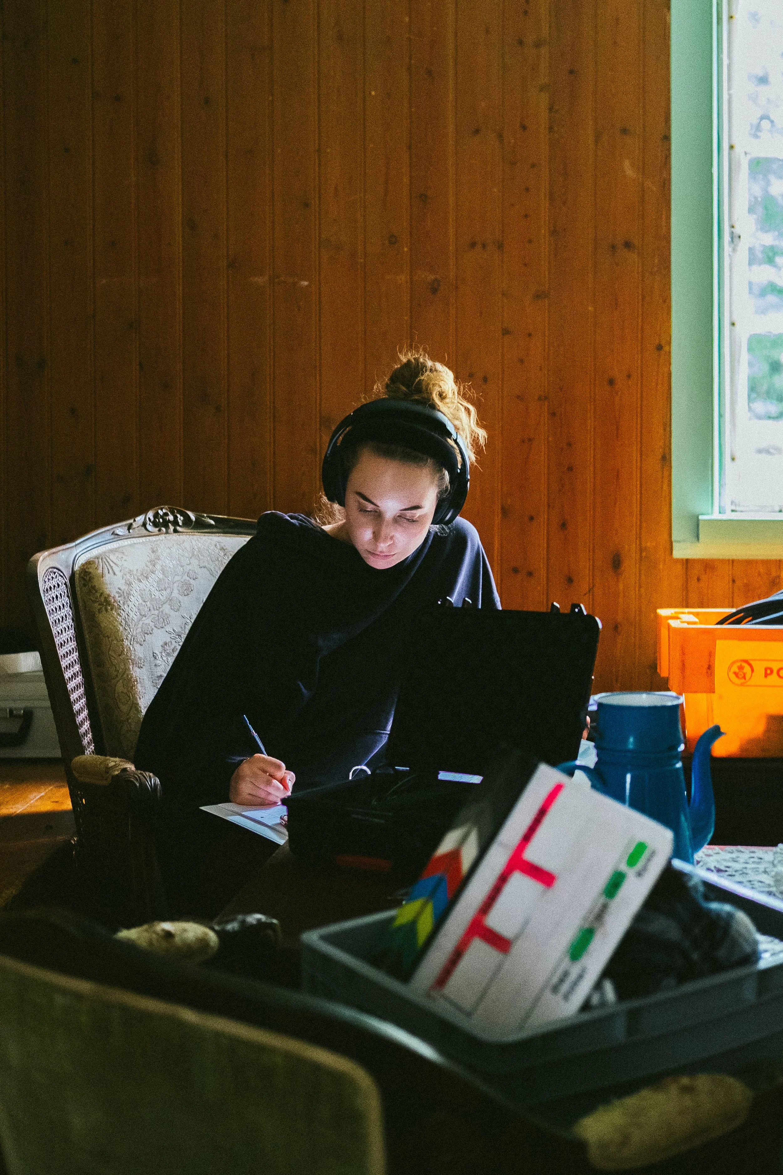 A young woman with curly hair wearing headphones, sitting at a table, writing on a notepad, with various objects including a colorful board game, a blue kettle, and an orange box nearby.