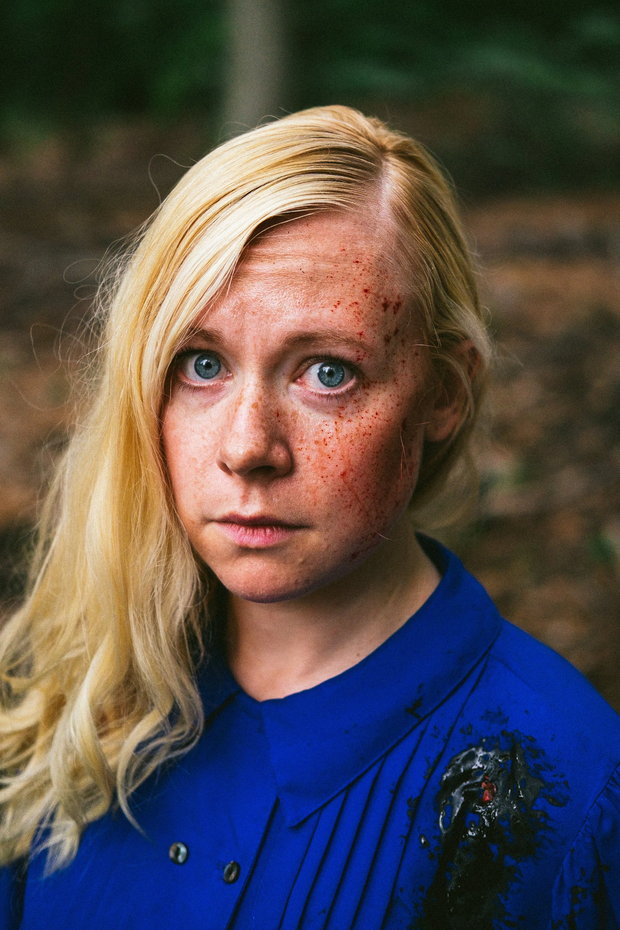 Close-up of a blonde woman with blue eyes, freckles, and some facial redness, wearing a blue shirt with a painted black and red design on the shoulder, outdoors with blurred greenery background.