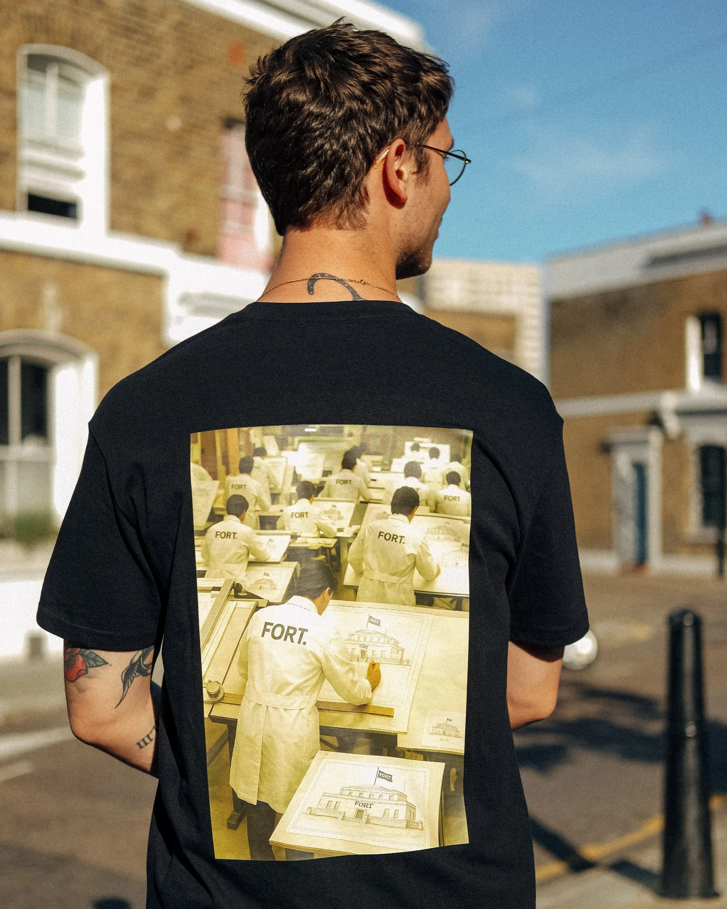 Rear view of a young man with brown hair, glasses, and a tattoo on his arm, standing outdoors with a clear blue sky and residential buildings in the background. He is wearing a black T-shirt with a graphic of people in yellow uniforms with 'FORT' wri