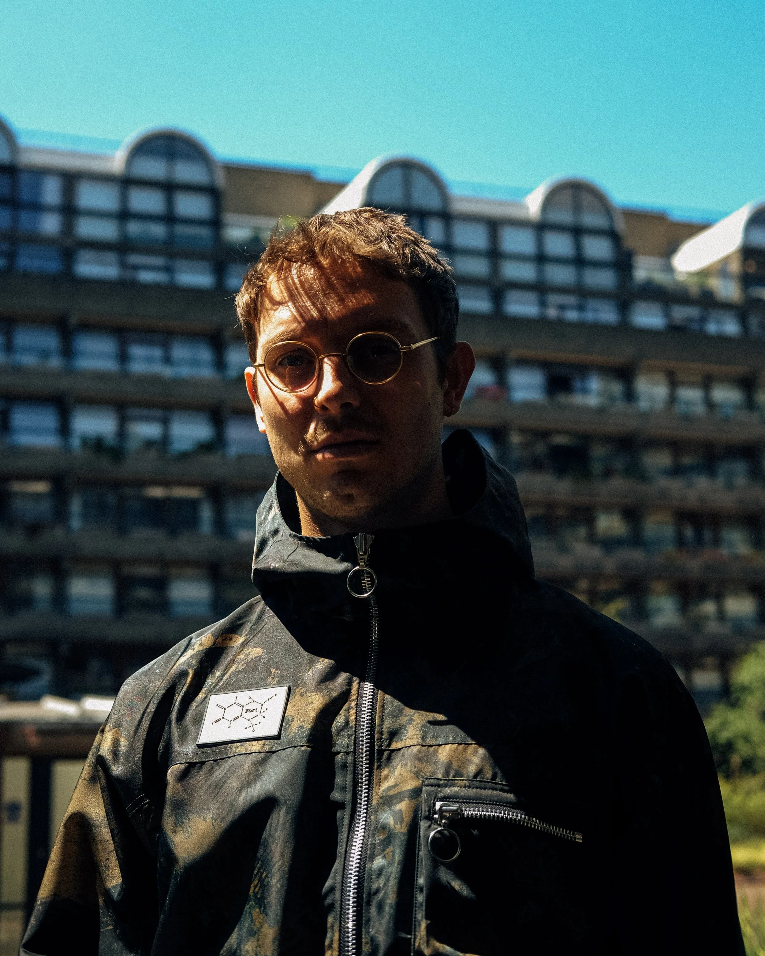 A young man wearing glasses and a camo jacket standing outdoors with a building and blue sky in the background.