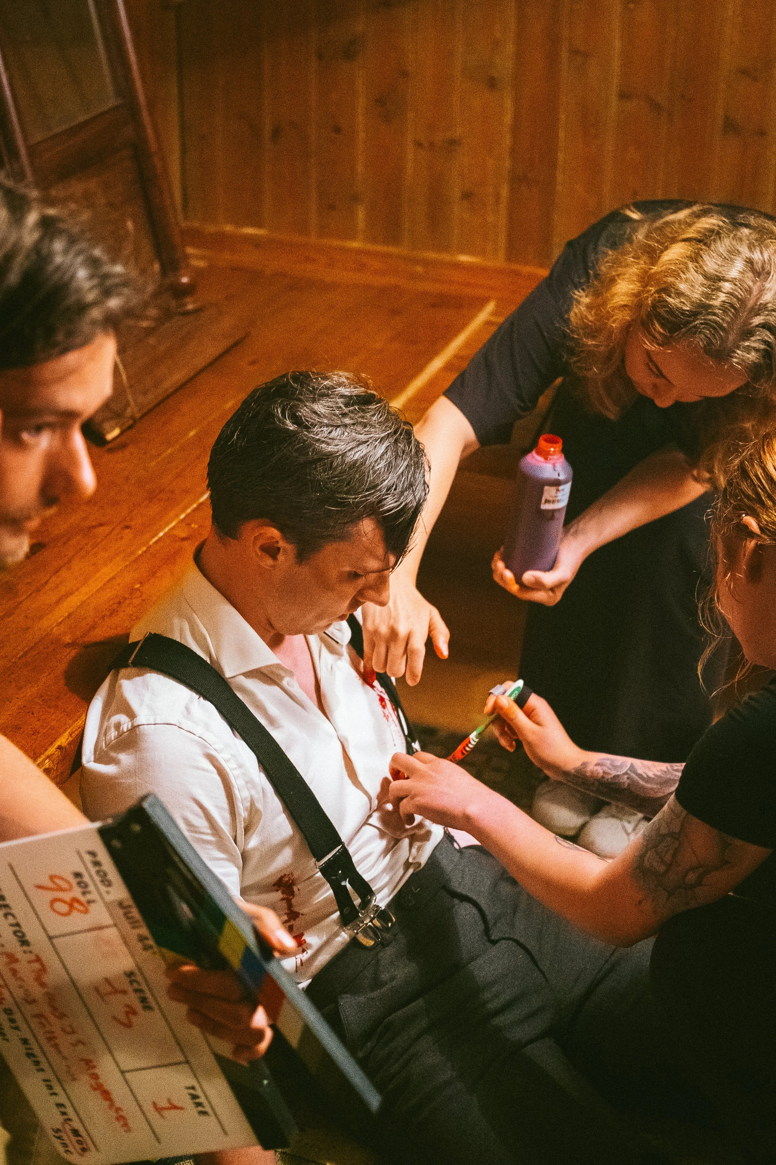 A woman with long curly hair, wearing black, is giving an injection or drawing blood from a seated man with dark hair, dressed in a white shirt and suspenders. There are two other individuals, one holding a bottle, and another with tattoos, assisting