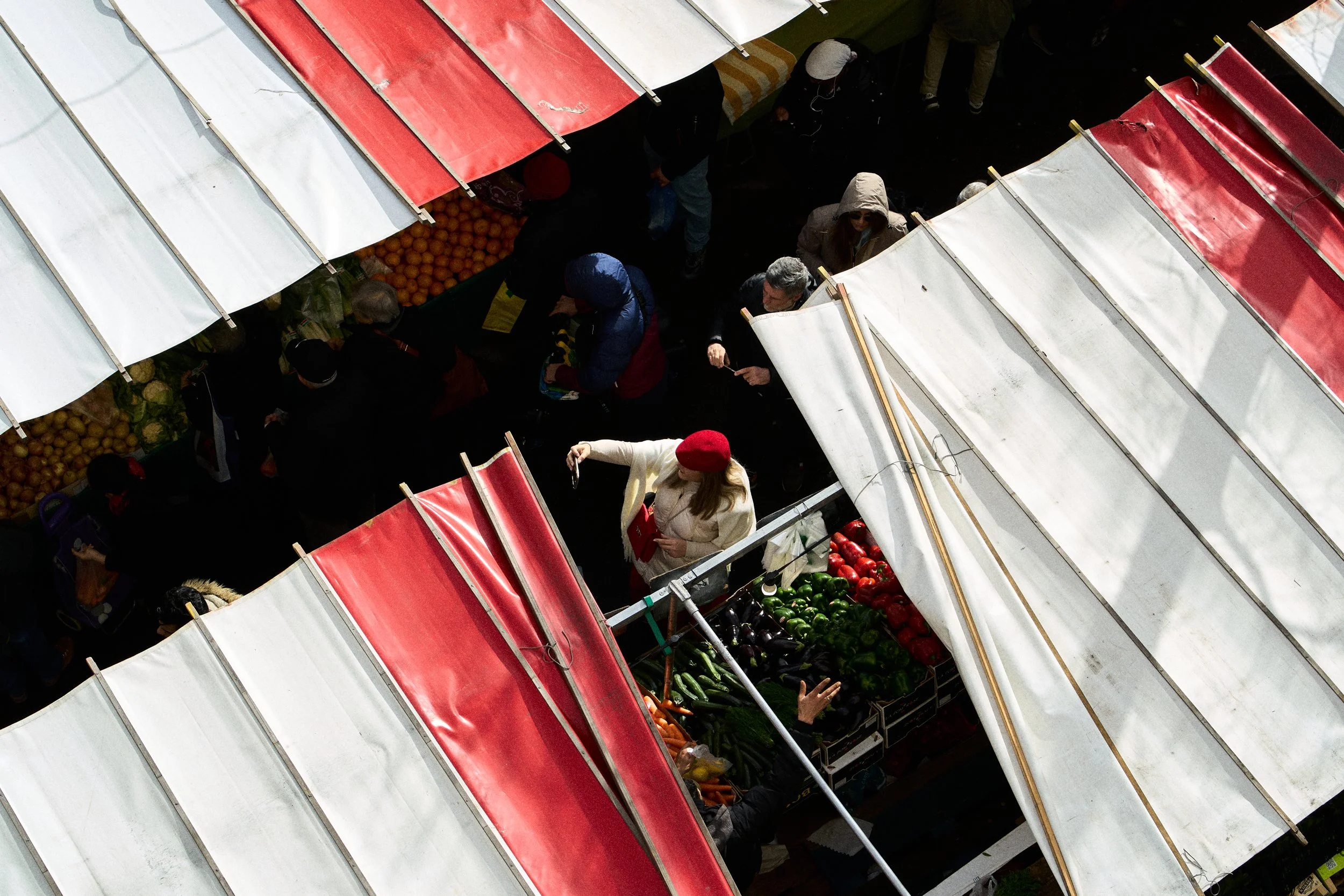 Overhead view of a busy outdoor market with vendors and shoppers browsing fruit and vegetable stalls under white and red canopies.