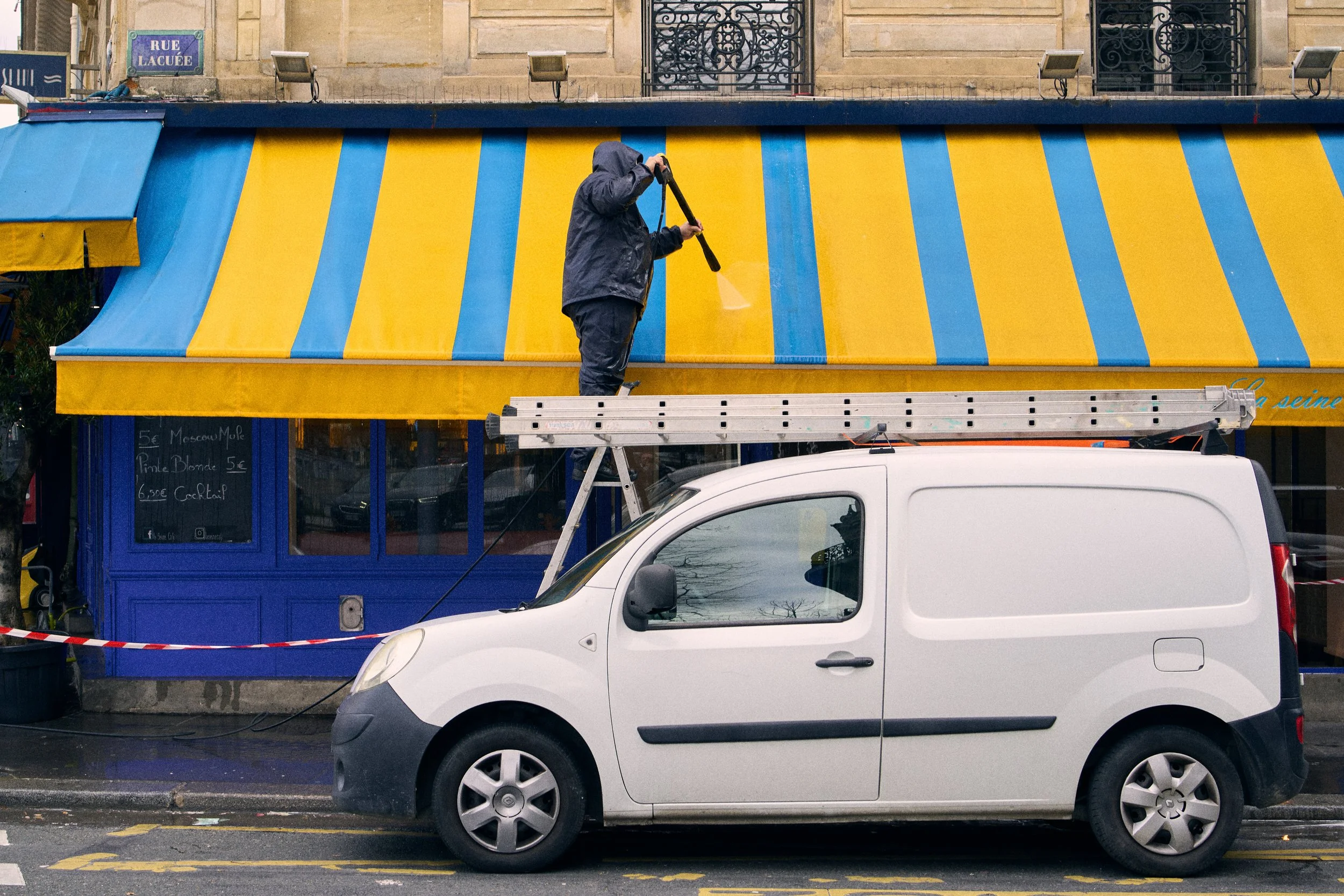 A person wearing a dark rain jacket and hood is standing on a tall ladder, cleaning the yellow and blue striped awning of a storefront. A white van is parked below, blocking part of the view of the storefront, which has French street signs on the bui