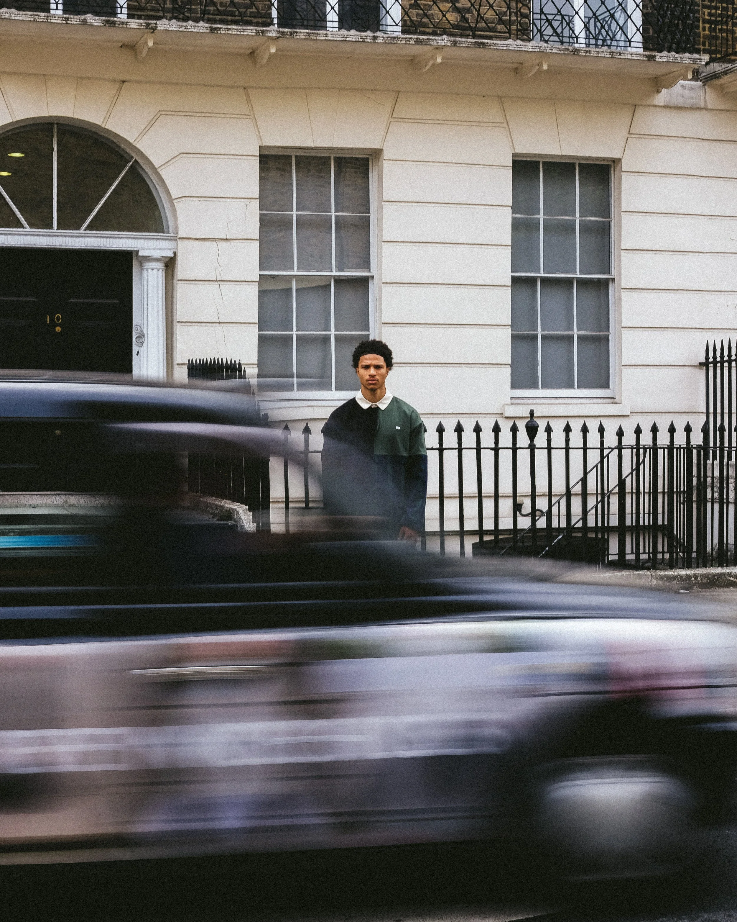 A young man with curly hair standing in front of a white building with two large windows, black wrought-iron fence, and a black car passing in the foreground.