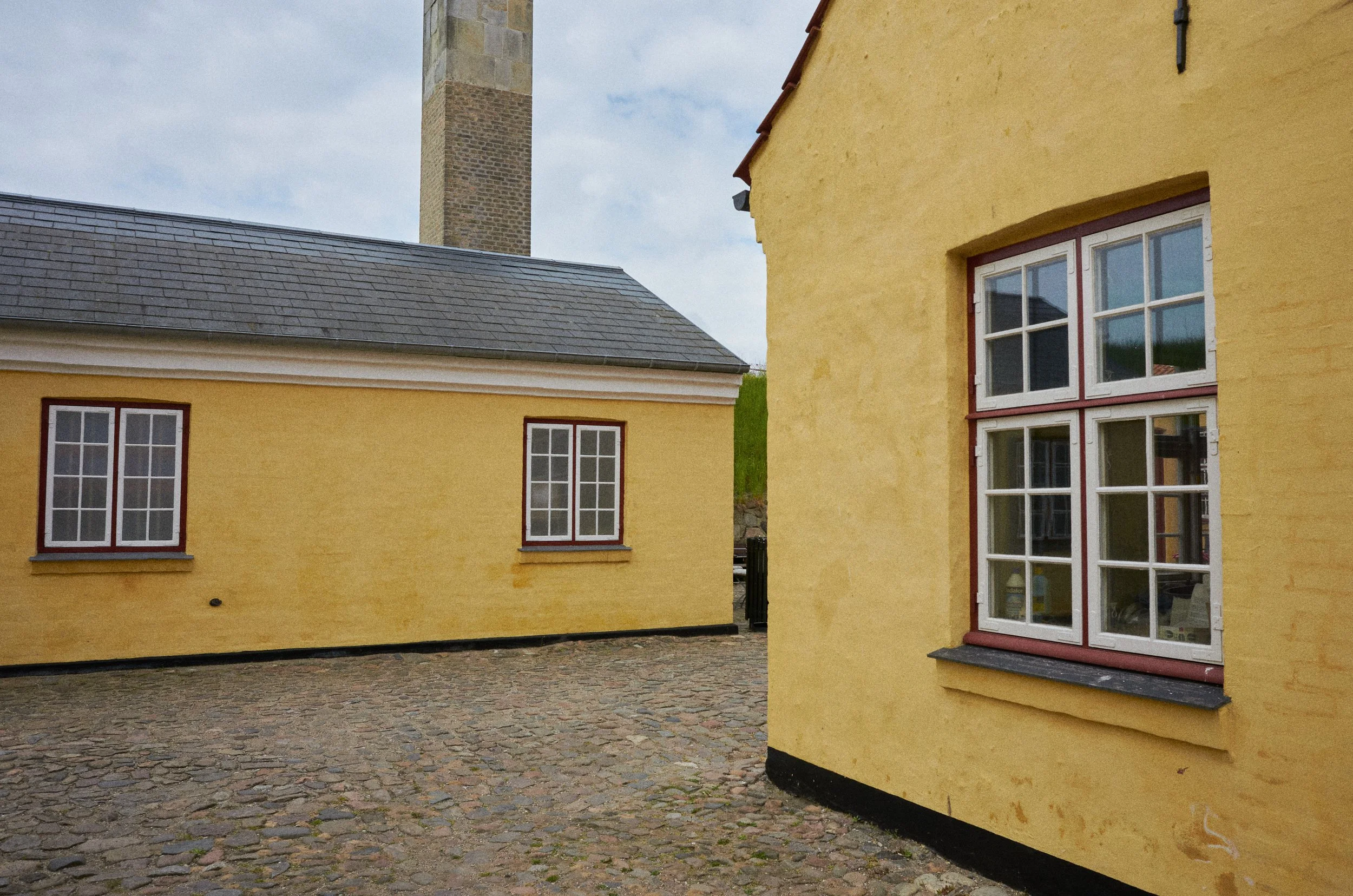 Yellow buildings with white windows and black foundations; cobblestone ground; stone chimney; cloudy sky.