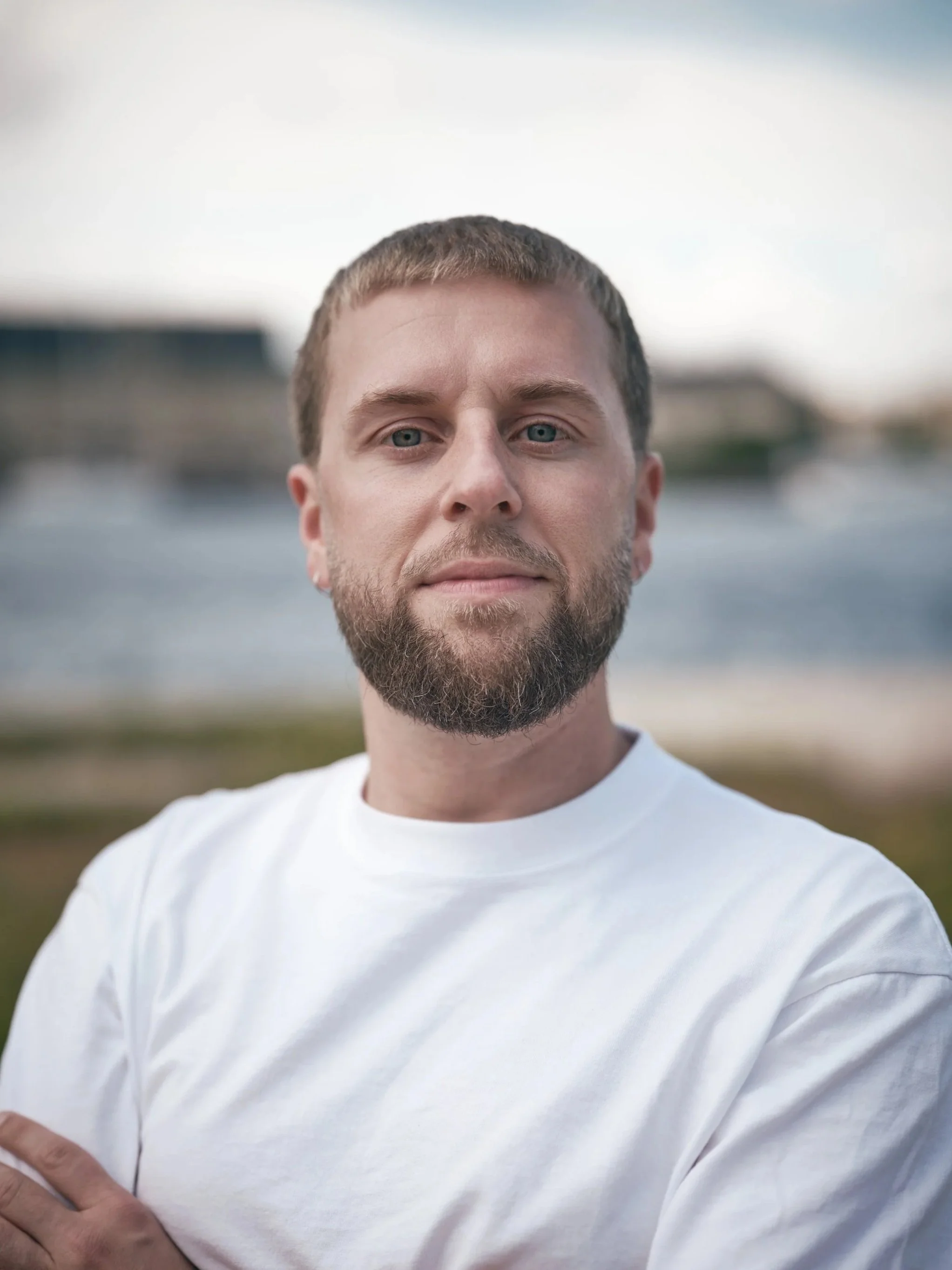 Close-up of a man with short brown hair and a beard, wearing a white shirt, standing outdoors with blurred buildings and sky in the background.