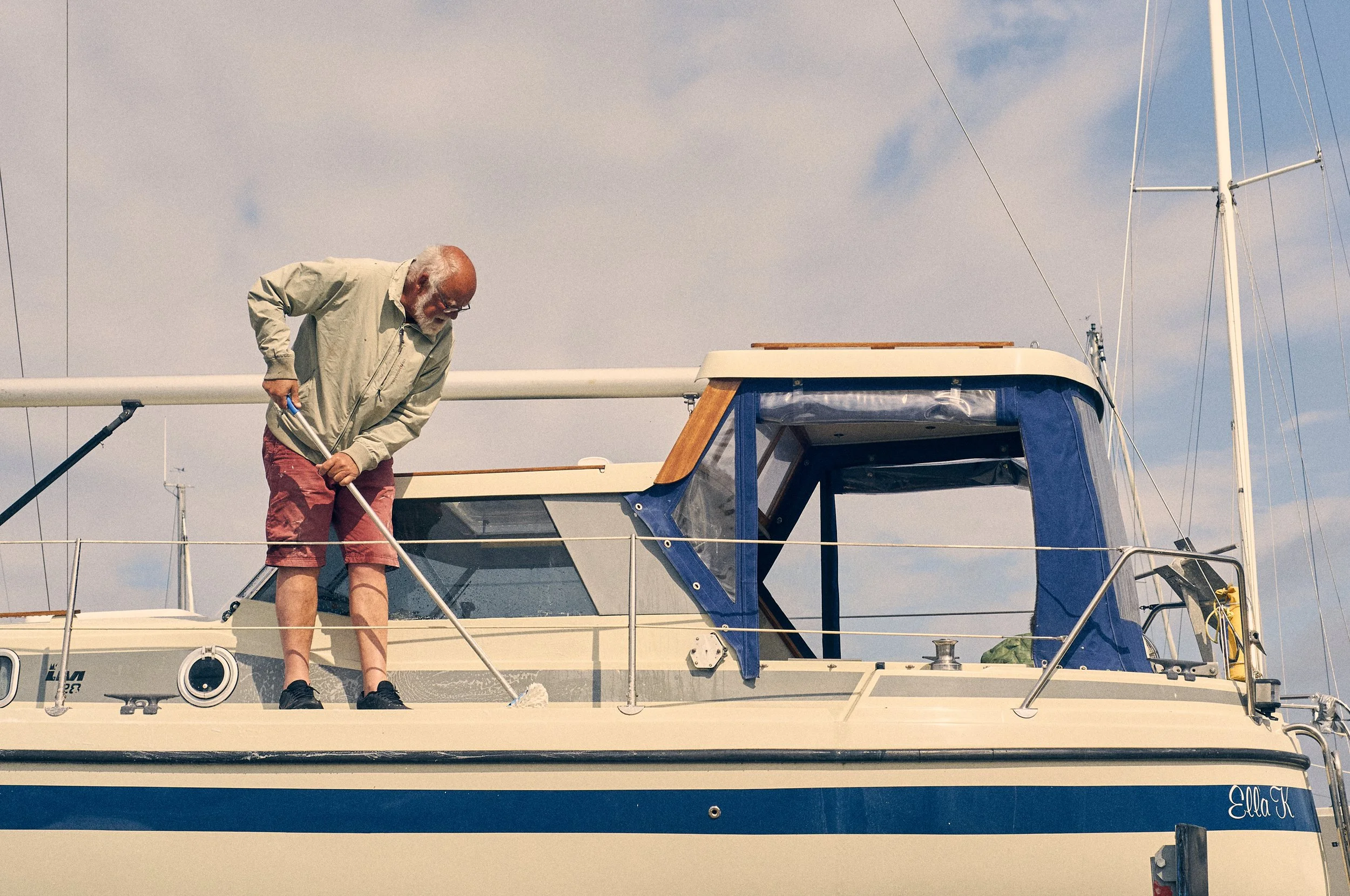 An elderly man with glasses, white hair, and a beard leaning over a sailboat's side, holding a cleaning tool, with sails and sky in the background.