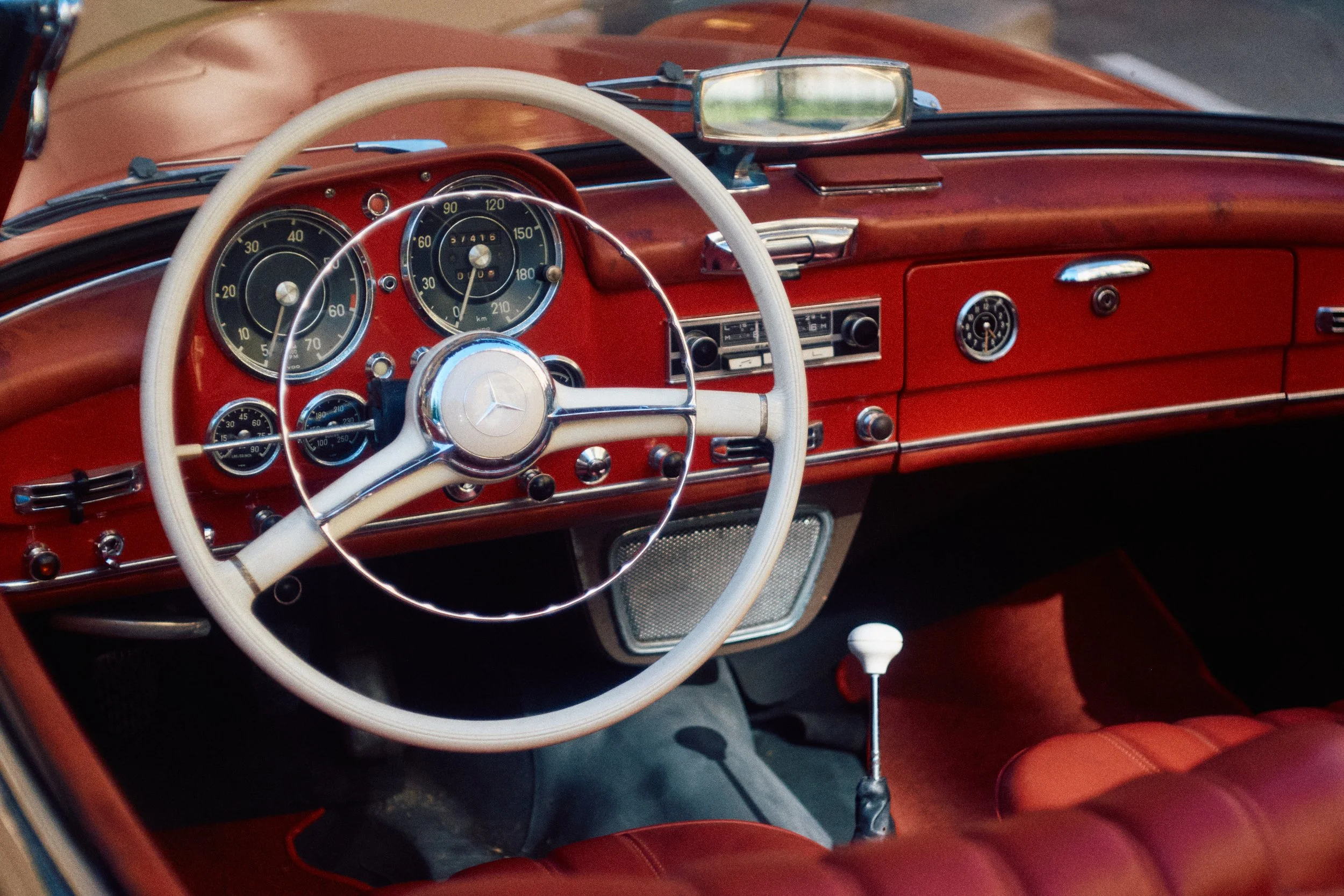 Interior of a vintage red car, showing the dashboard with analog gauges, a steering wheel with a Mercedes-Benz logo, and a gear shift. The dashboard features a radio, clock, and other controls.