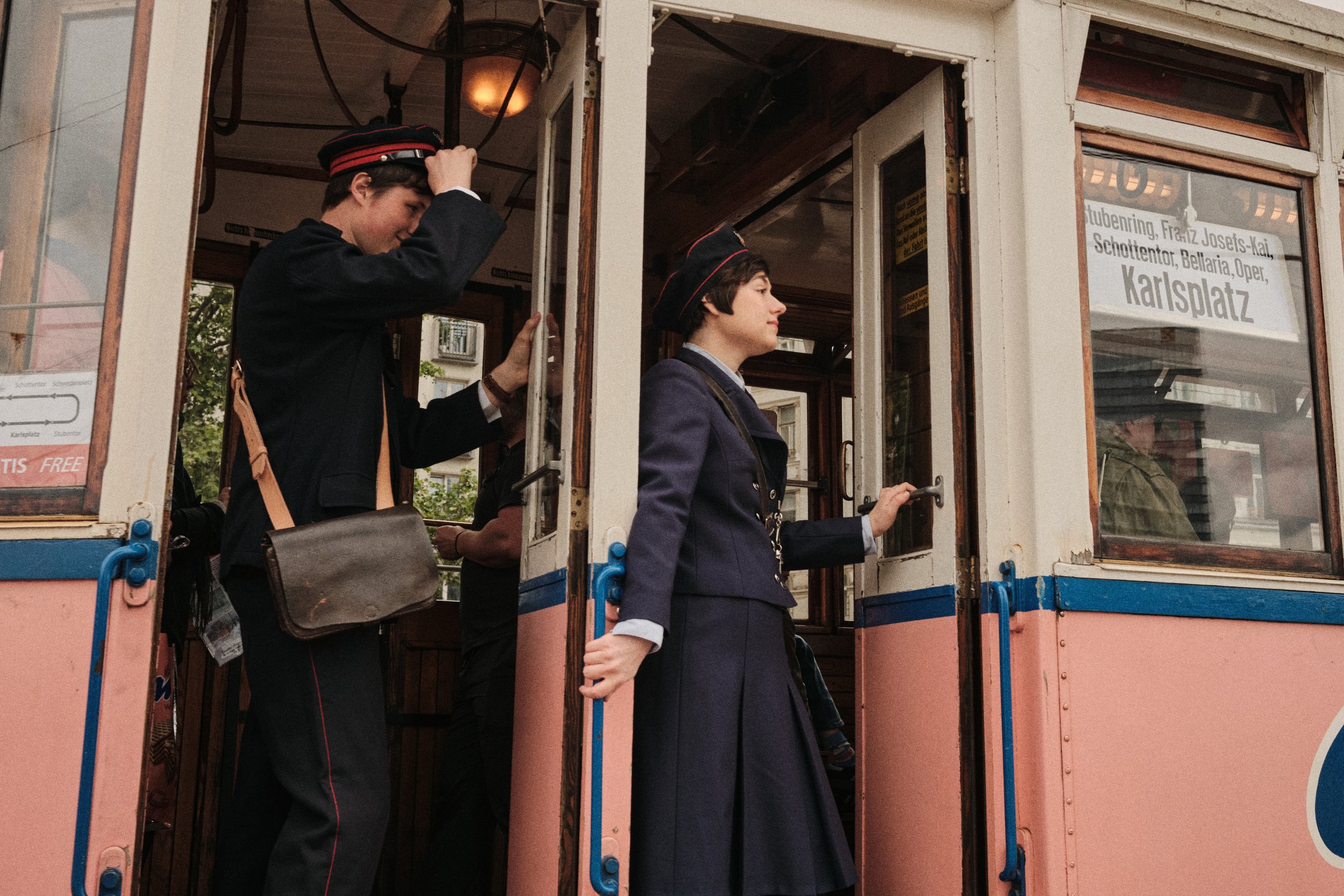 Two young women dressed as train conductors riding a vintage tram. One is standing at the door, holding the handle, and the other is adjusting her hat while looking down.