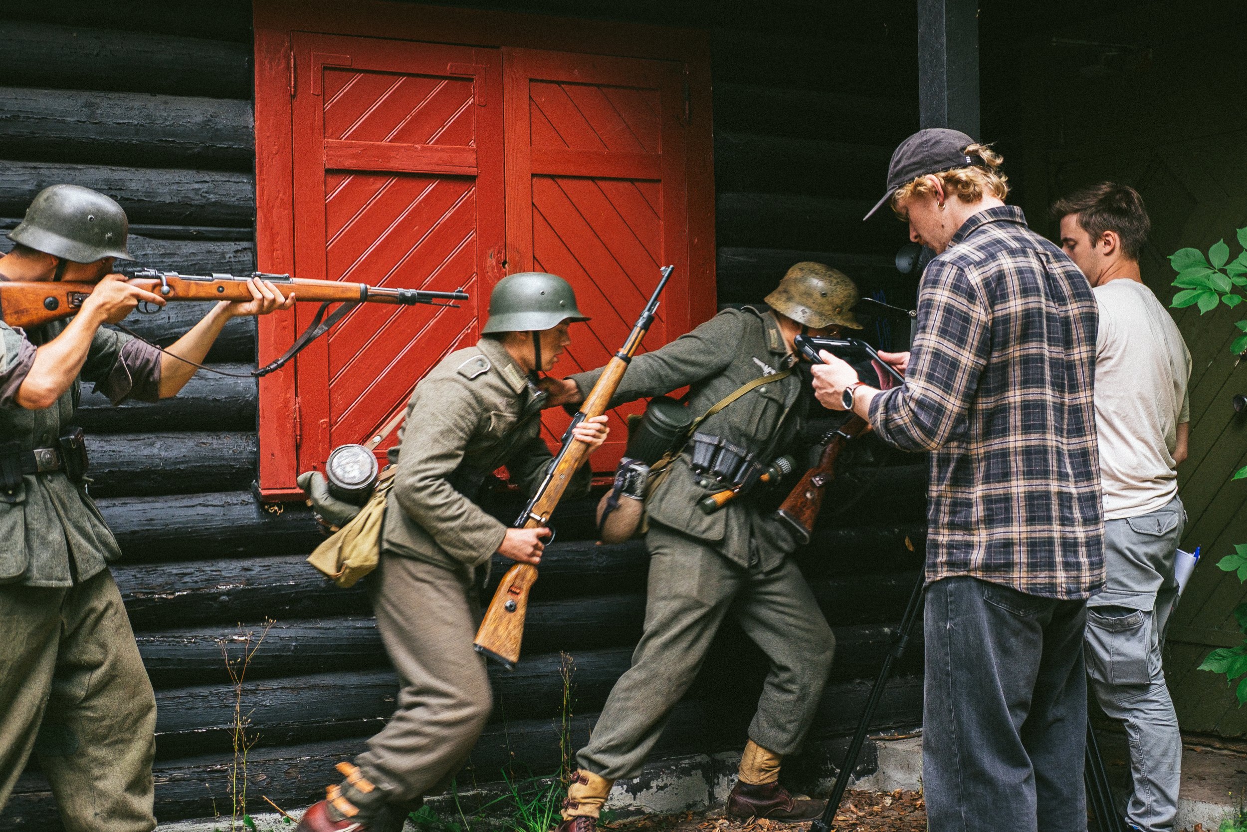 People dressed in military uniforms and a few civilians, some wearing helmets, moving in or around a black wooden building with a red door. The scene appears to be historical, possibly from a wartime reenactment or film set.
