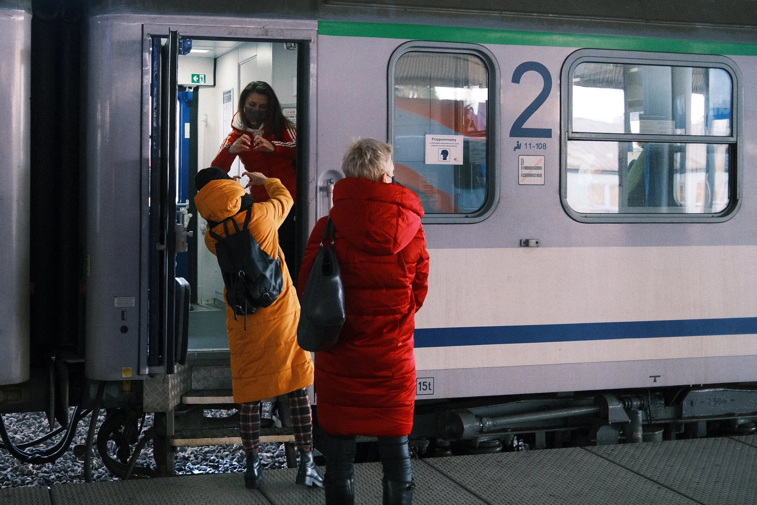 Two women and a child standing near a train, with a woman inside the train making a heart shape with her hands. All are wearing winter coats, and the women are wearing face masks.