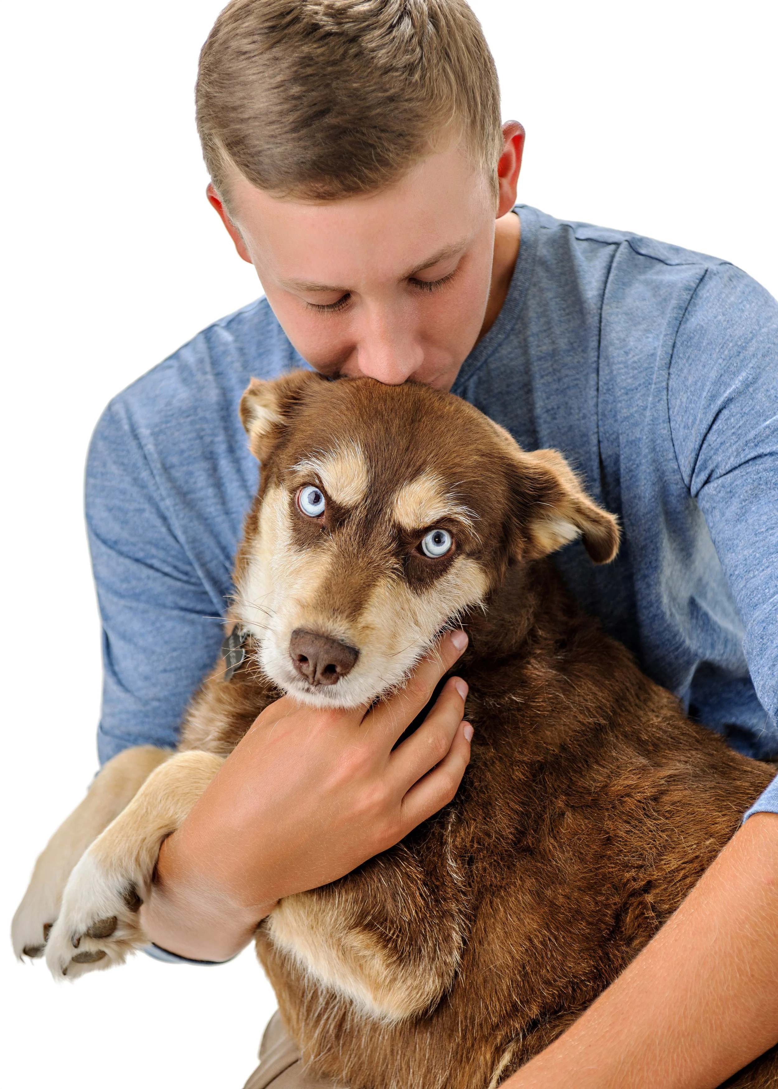 A young man with closed eyes gently kisses a brown and white Siberian Husky puppy with piercing blue eyes, both holding each other affectionately.