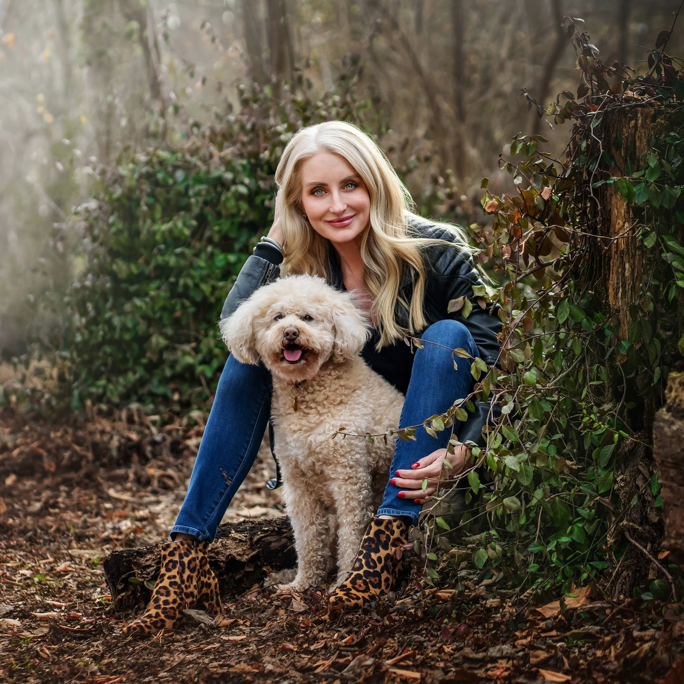 A blonde woman in a black jacket and leopard print boots sitting outdoors in a wooded area with a fluffy dog.